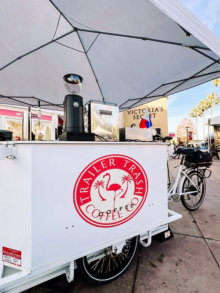 A mobile coffee cart with the logo 'Trailer Trash Coffee' featuring a pink flamingo, palm trees, and a red circle, parked outdoors under a large umbrella, with a bicycle attached to it. In the background, there's a Victoria's Secret store and some pedestrians.