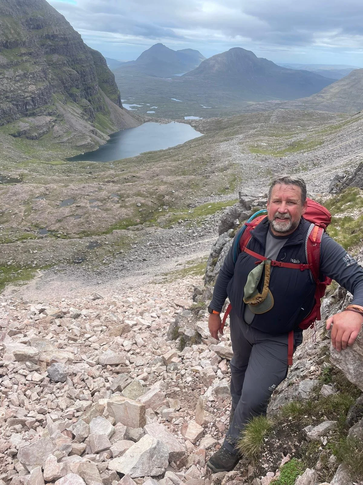 A man hiking on a rocky mountain trail next to a steep slope with a scenic view of a mountain lake and distant mountains under cloudy skies.