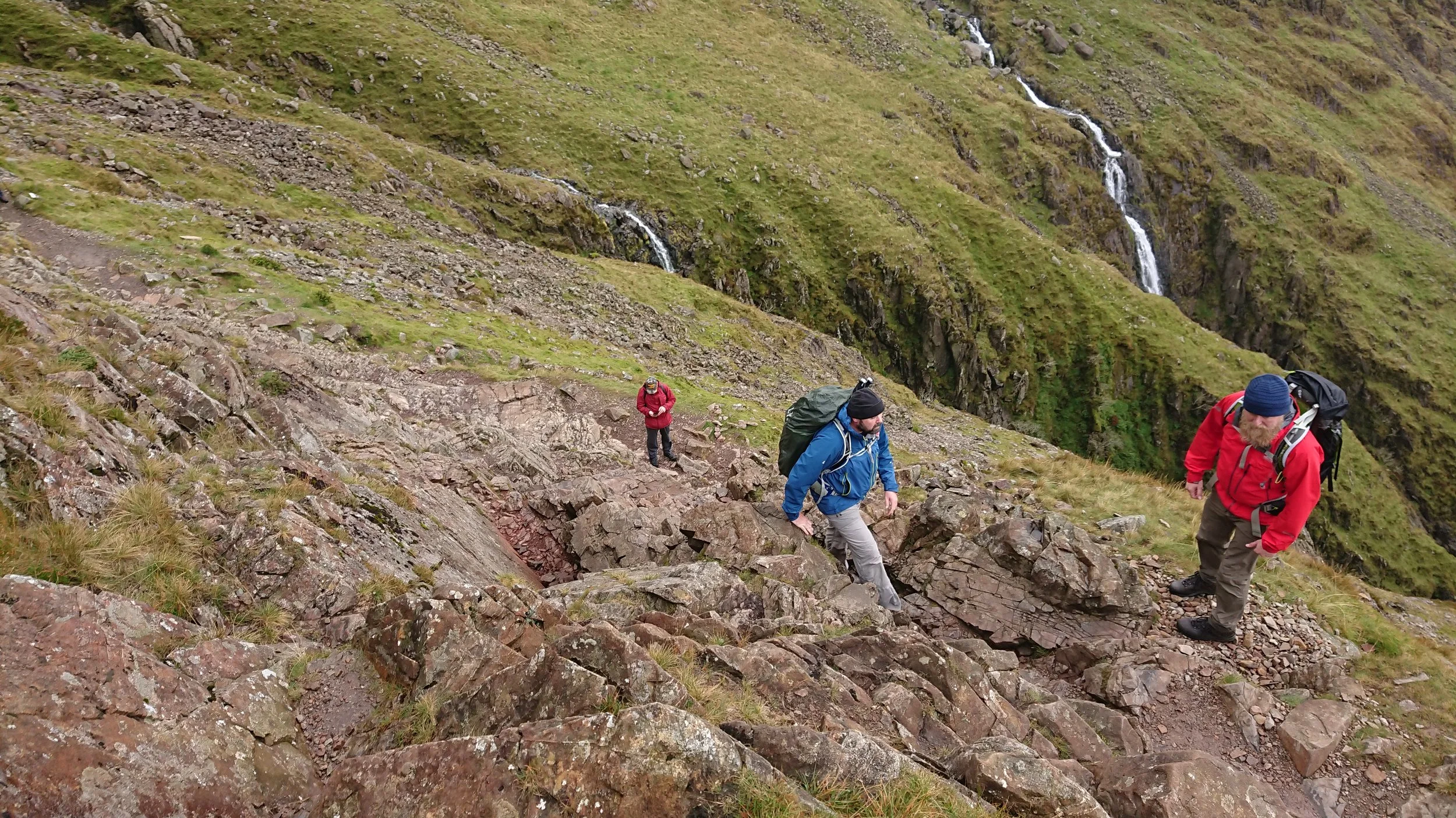 Three hikers, two men and one woman, wearing outdoor gear and backpacks, climbing a steep rocky mountain trail with patches of grass. A small waterfall flows down the green, rocky mountainside in the background.