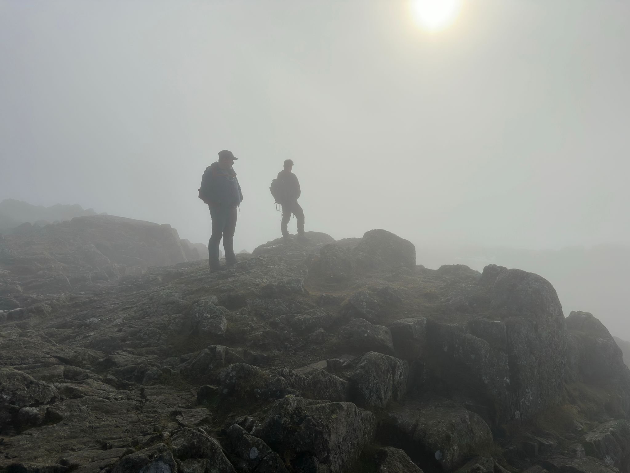 Two hikers stand on rocky terrain amidst foggy conditions with the sun faintly visible through the haze, creating a mysterious mountain scene.