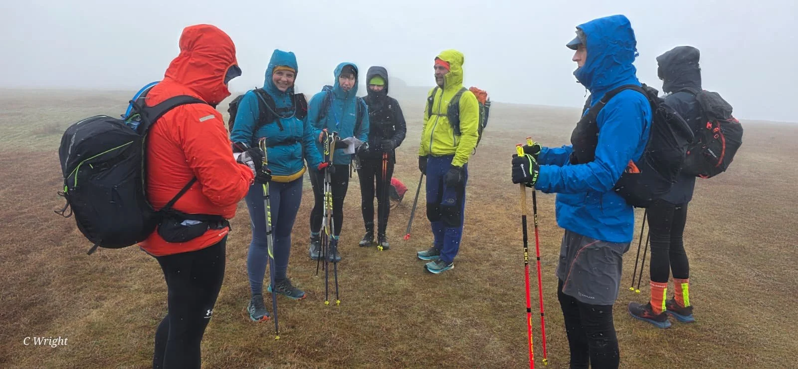 Group of six hikers in outdoor gear standing on a foggy, grassy field, some holding trekking poles and wearing backpacks.
