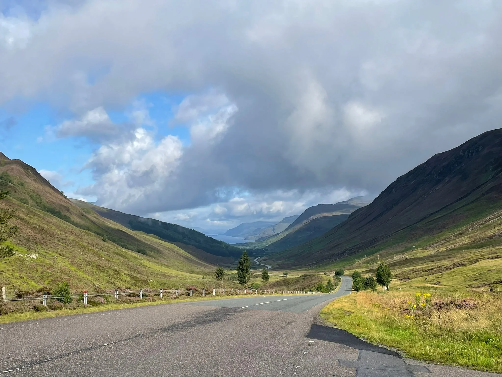 A winding road through a valley surrounded by green hills and mountains under a partly cloudy sky.