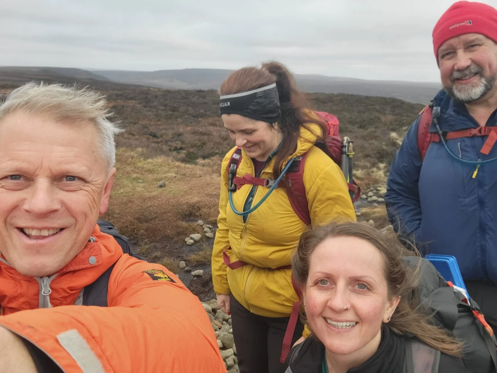 A group of five smiling hikers taking a selfie outdoors on a cloudy day with a rocky and grassy landscape in the background.
