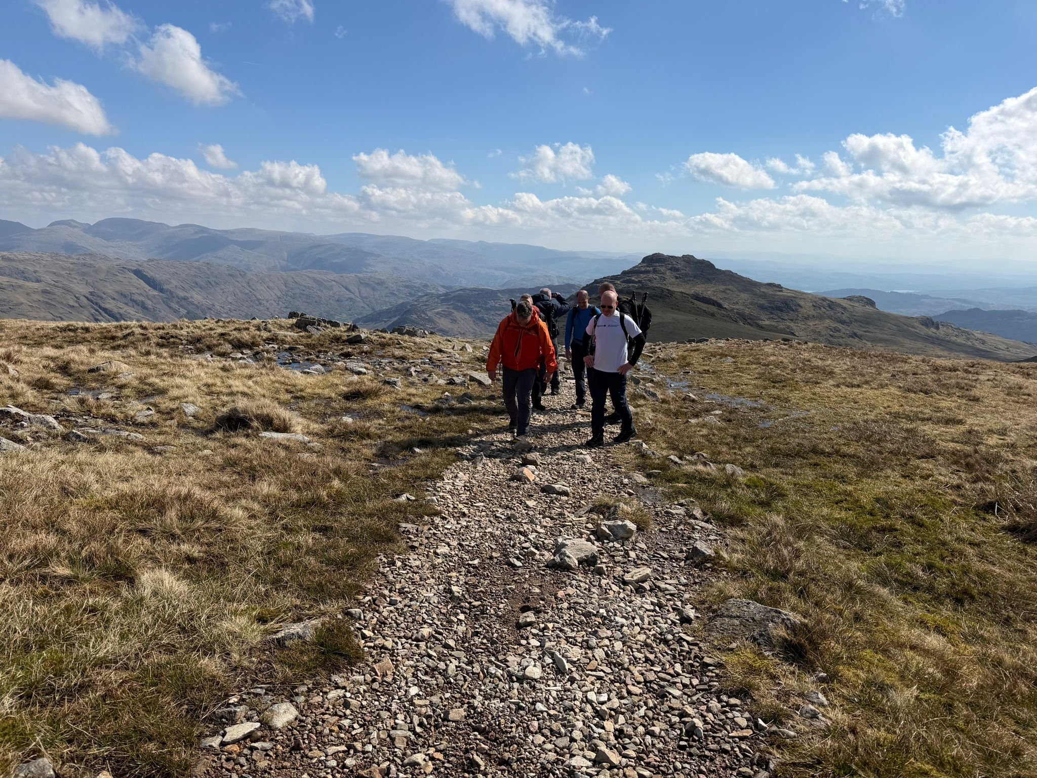A group of five hikers walking along a rocky trail in a mountainous landscape under a partly cloudy sky.