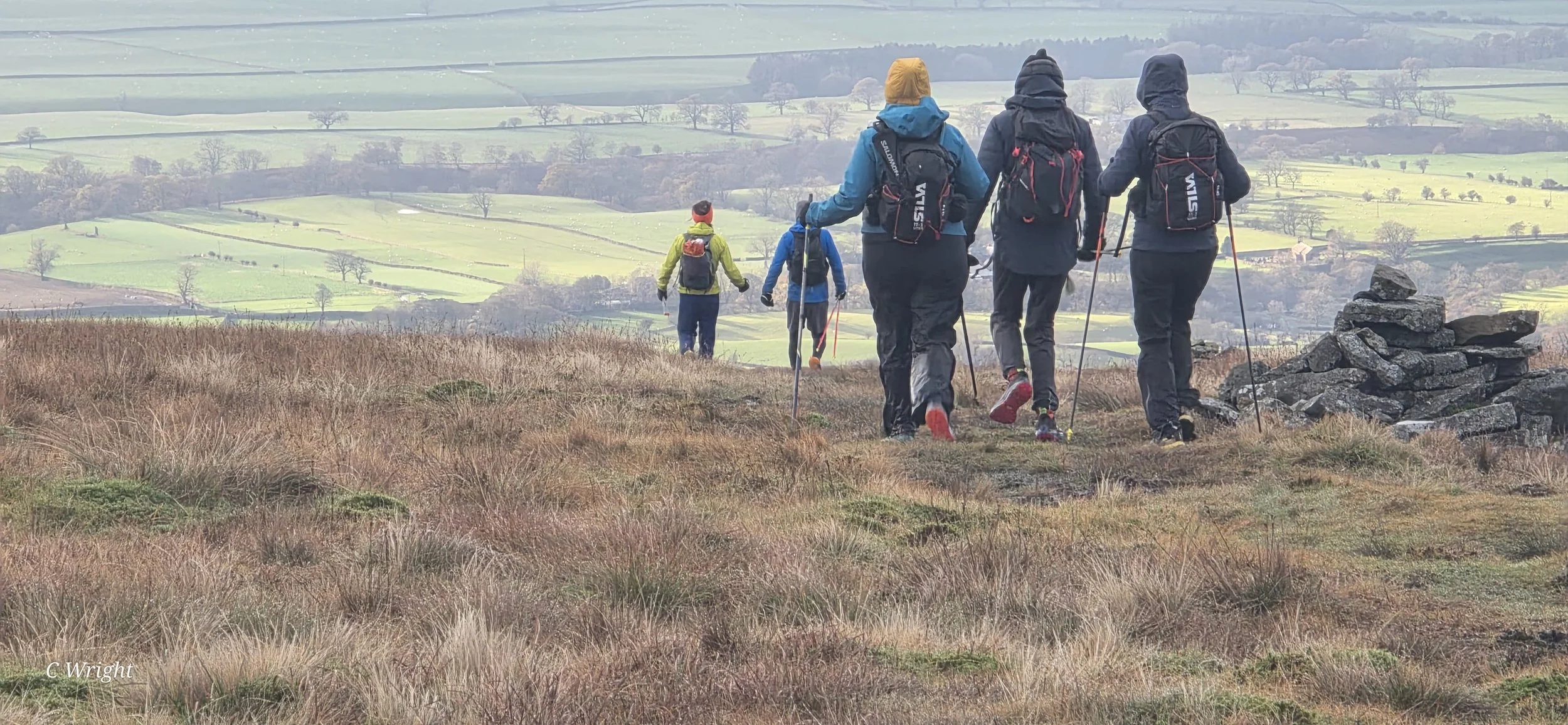 A group of hikers walking on a grassy hill with rolling fields and trees in the background, wearing rain jackets and backpacks.