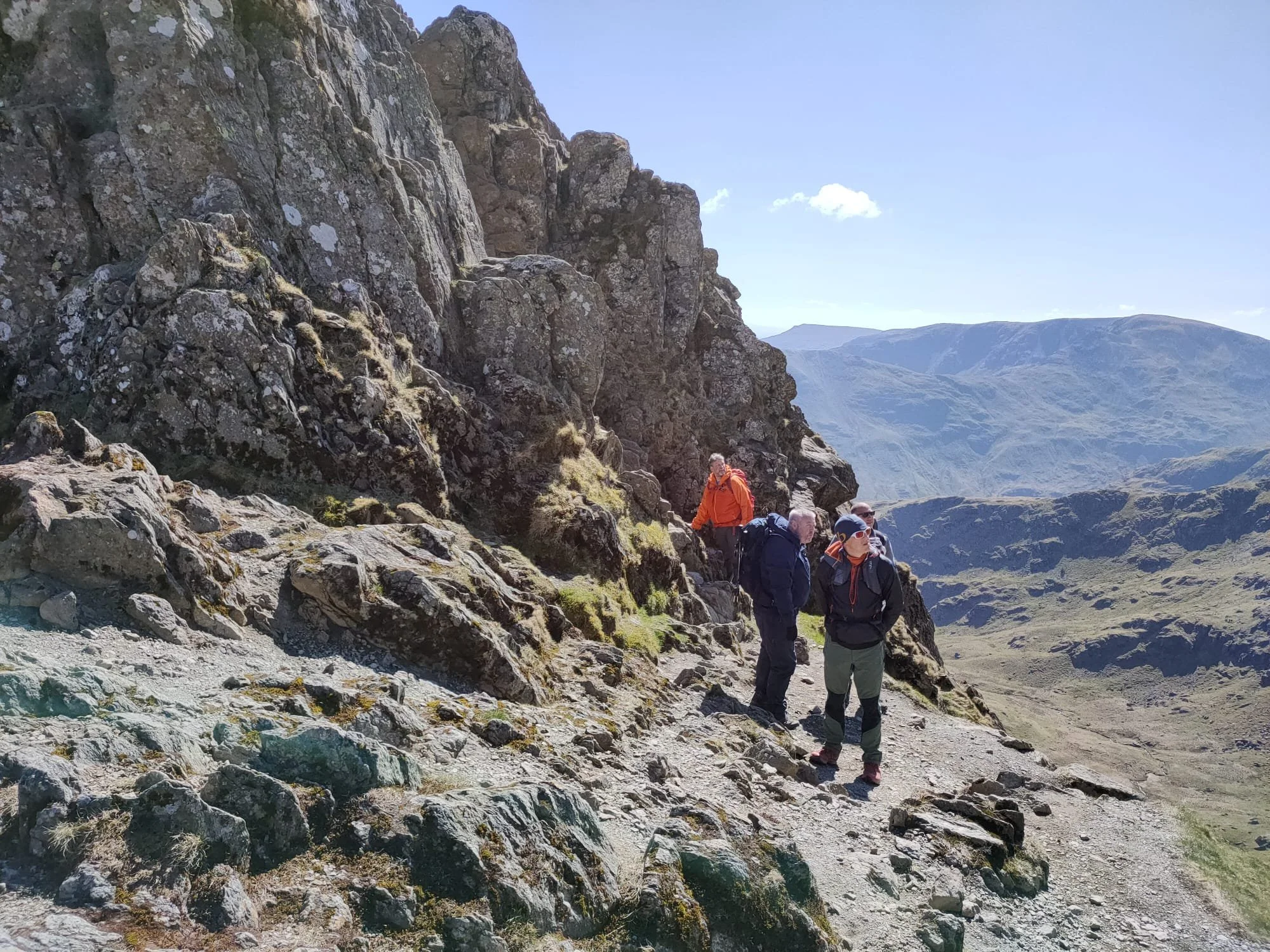 A group of hikers ascending a rocky mountain trail with a steep cliff on one side and expansive mountain ranges in the background under a clear blue sky.