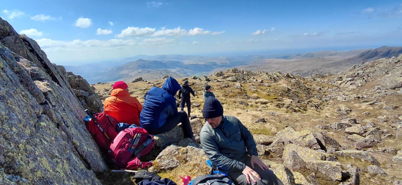 Hikers resting on rocky mountain landscape with expansive valley and distant mountains under a partly cloudy sky.