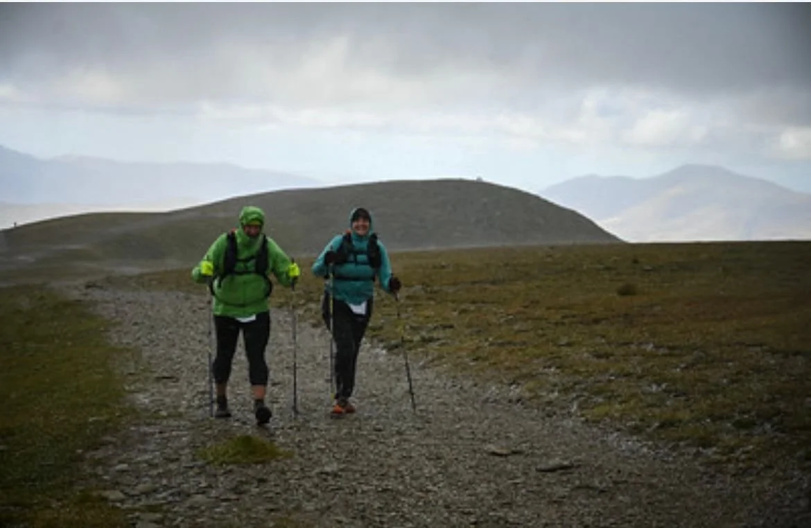 Two hikers walking on a rocky trail in a mountain landscape with overcast skies.