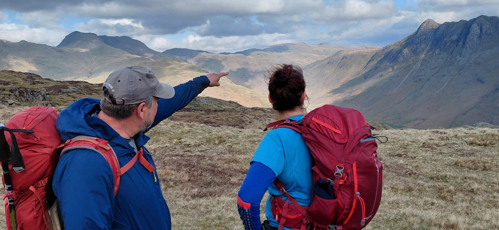 Two hikers in outdoor gear, backpacks, and hats, standing on a grassy hillside, looking at a mountain range, one pointing toward the distance.