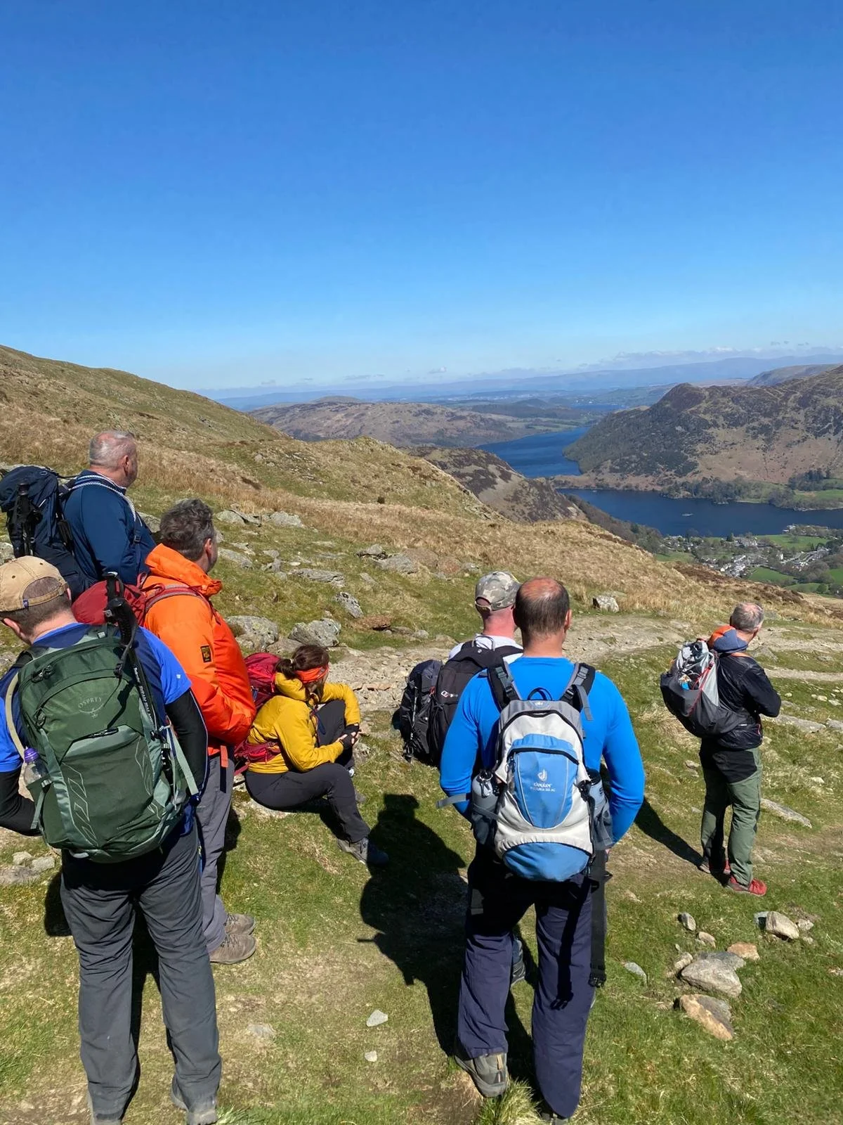 Group of hikers resting on a grassy hillside overlooking a scenic lake and mountains under a clear blue sky.