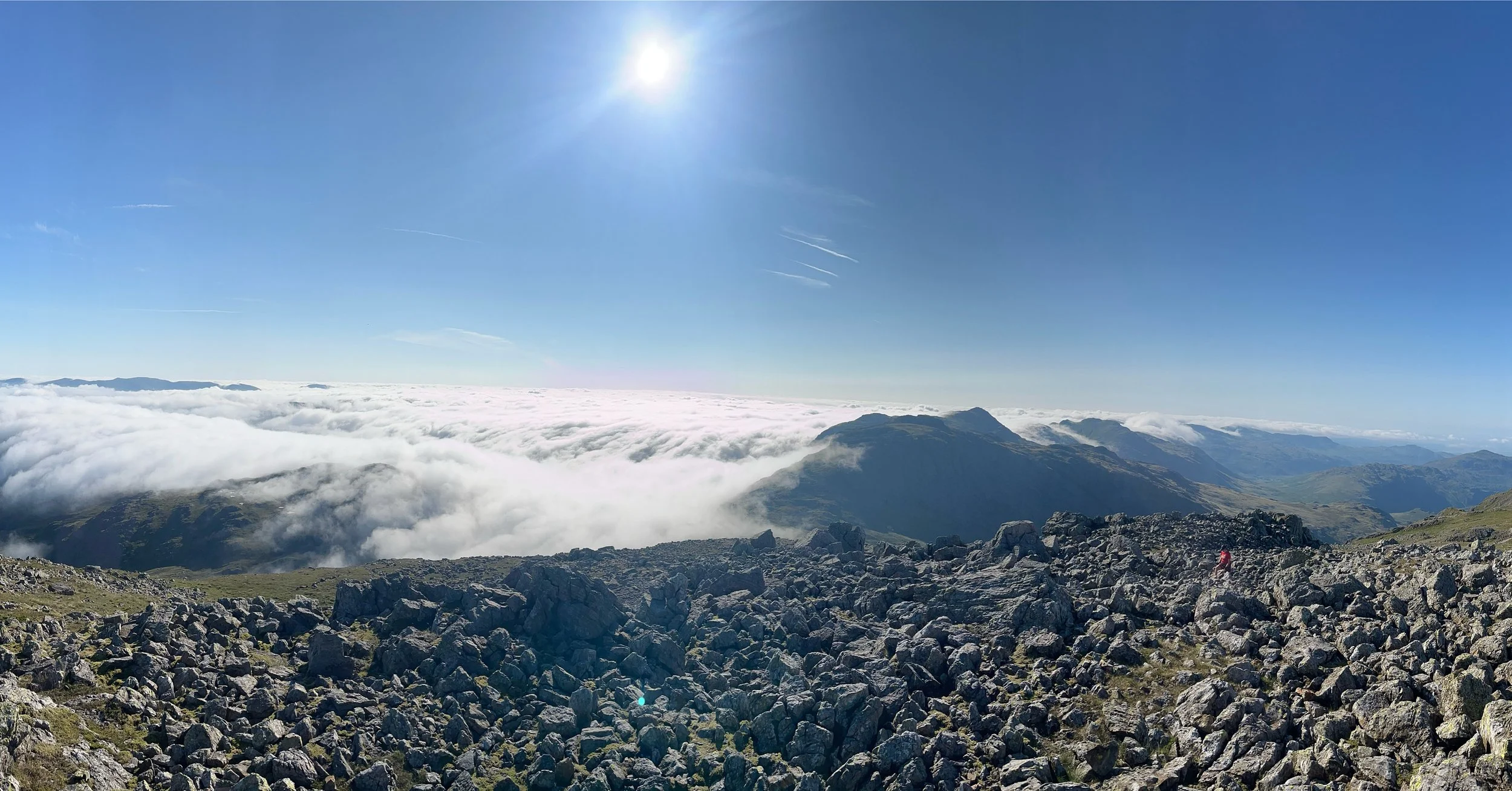 A scenic view from a mountain summit showing a rocky terrain in the foreground, a layer of clouds below, distant mountain ridges, and a bright sun high in a clear blue sky.