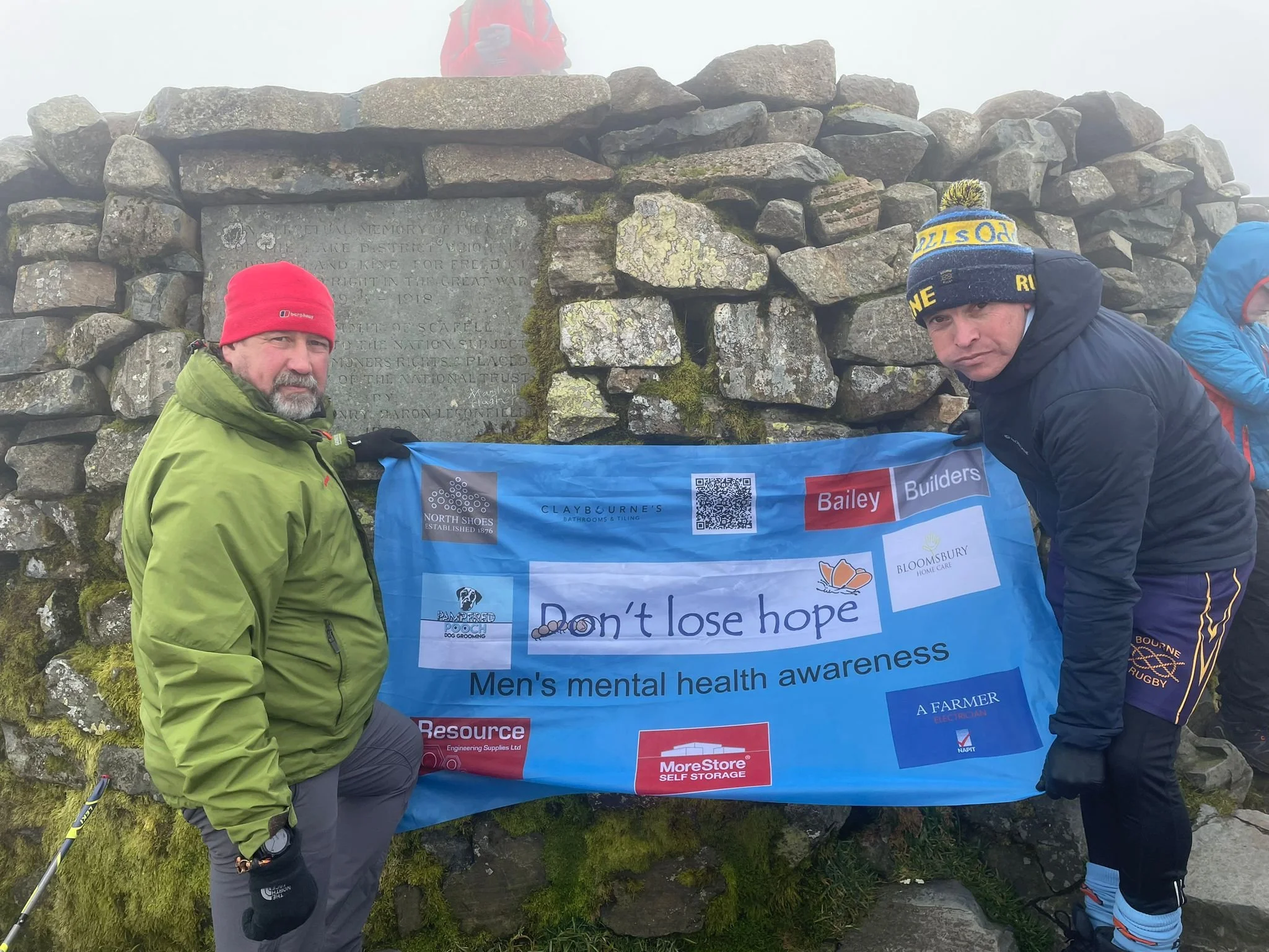 Three men in outdoor clothing holding a blue banner with various sponsor logos and the message "Don't lose hope" for men's mental health awareness, standing in front of a stone monument.