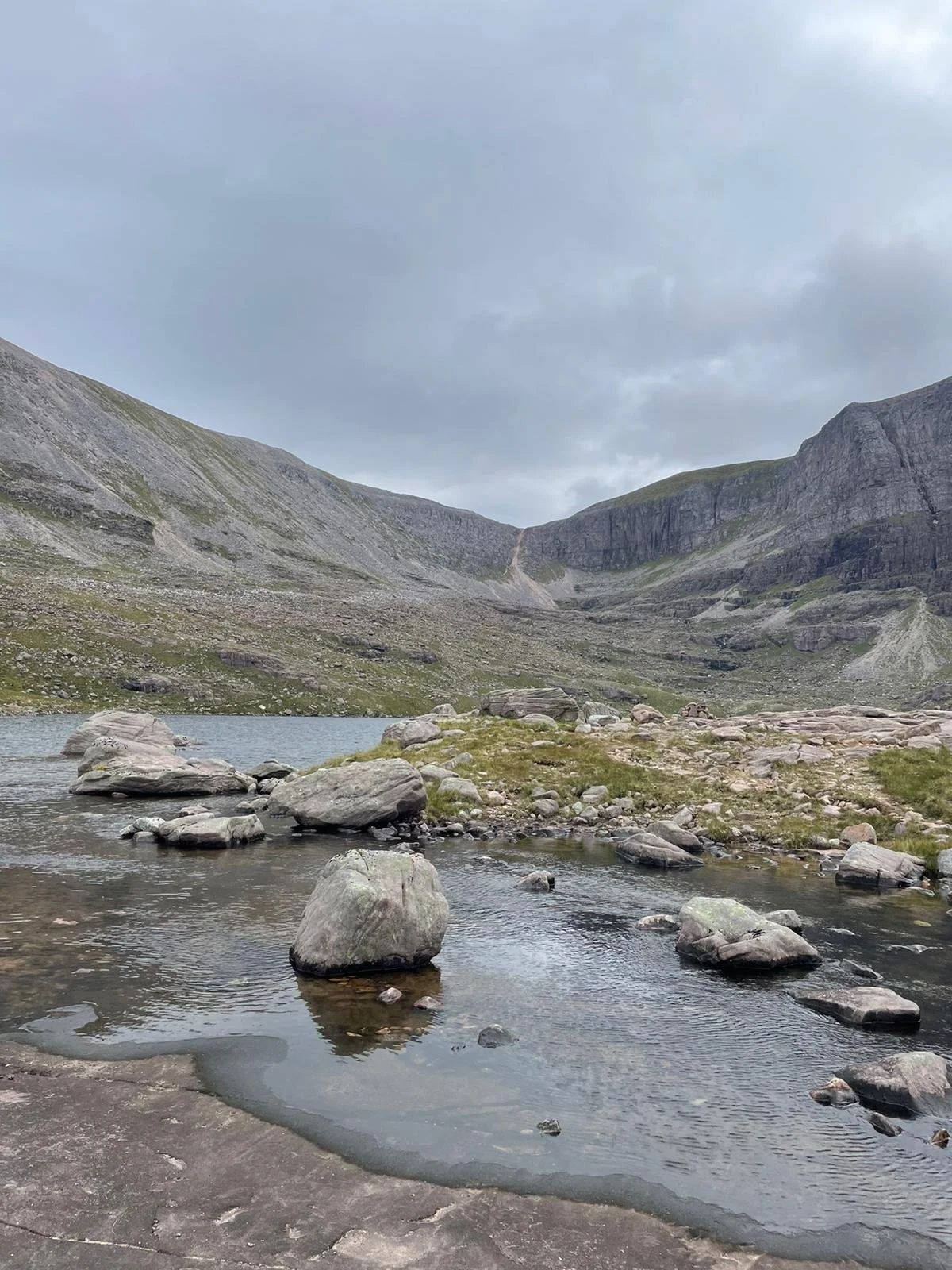A mountain landscape with a small lake and rocks in the foreground, surrounded by grassy and rocky slopes under a cloudy sky.