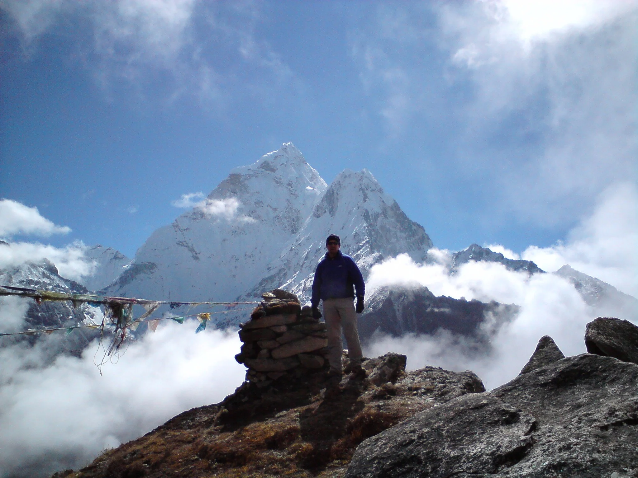 A person standing on a rocky ledge in front of a snow-capped mountain peak with a blue sky and some clouds.