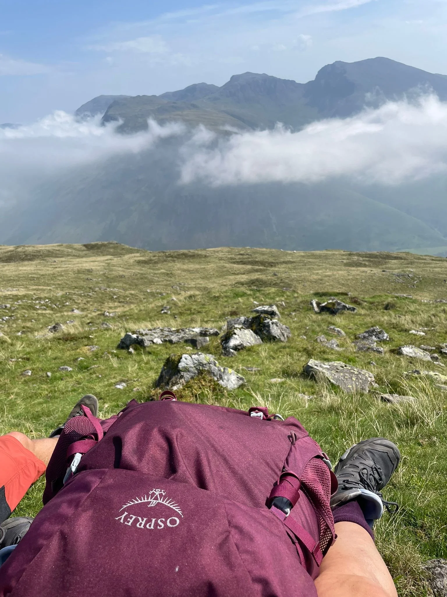 Person lying on a grassy hillside with a backpack, wearing hiking shoes, with a scenic view of mountains, clouds, and sky in the distance.