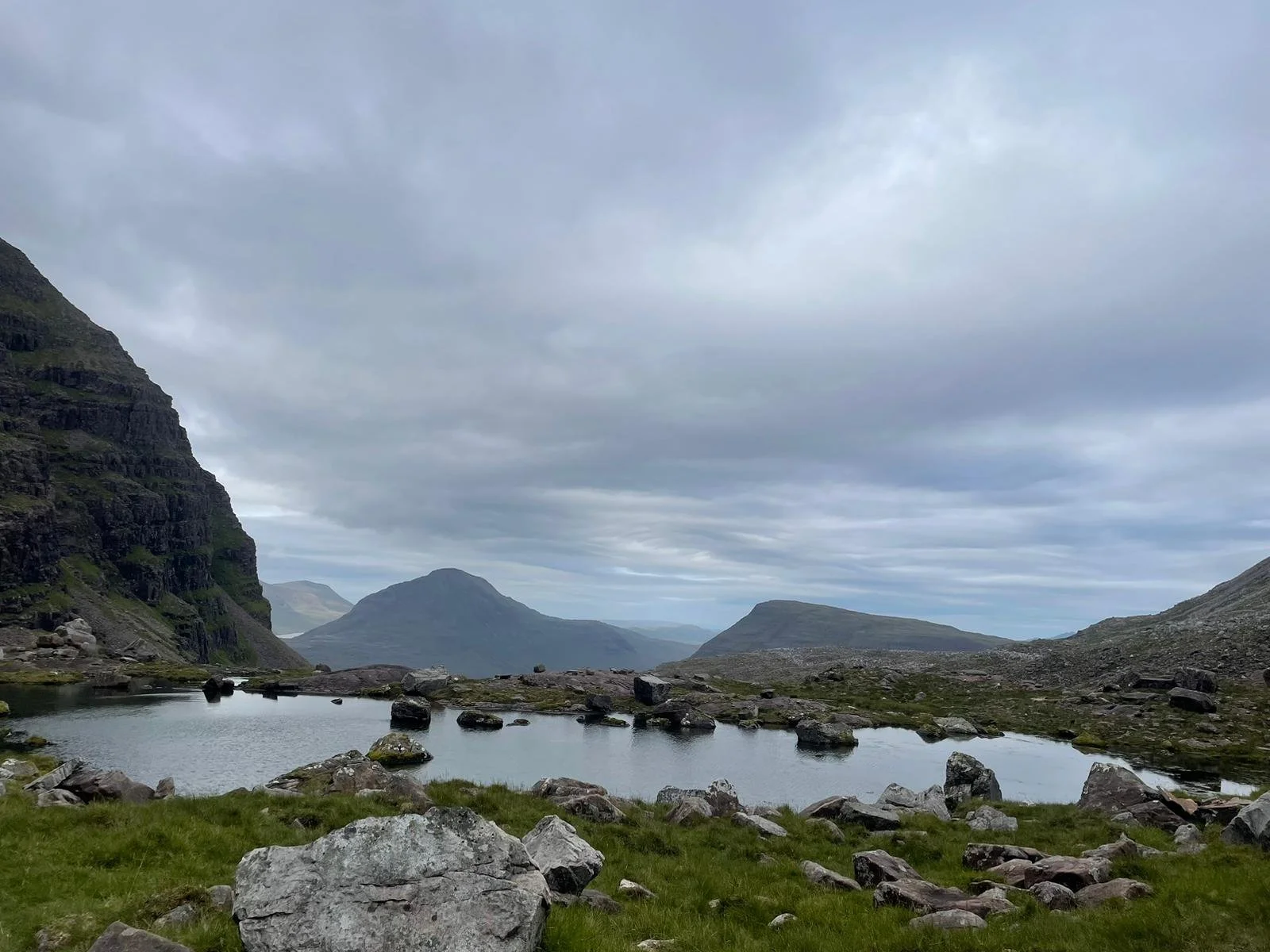 A mountainous landscape with a small pond in the foreground, surrounded by rocks and green grass, under a cloudy sky.