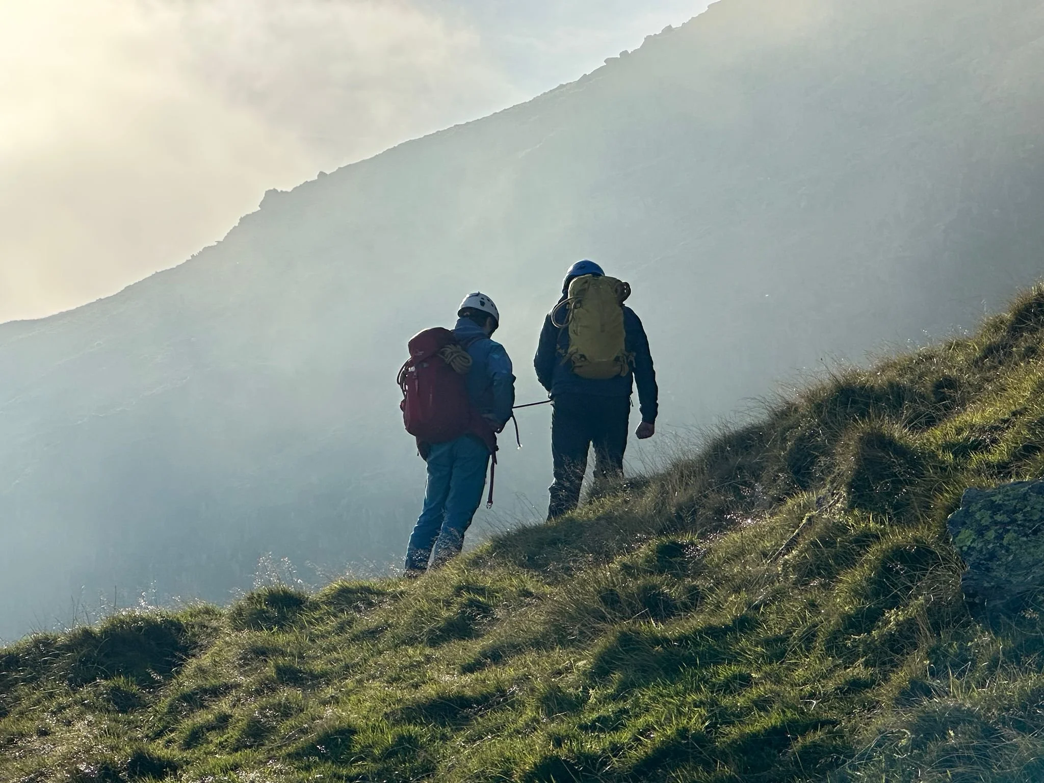 Two hikers with backpacks walking along a grassy mountain trail surrounded by mist and clouds.