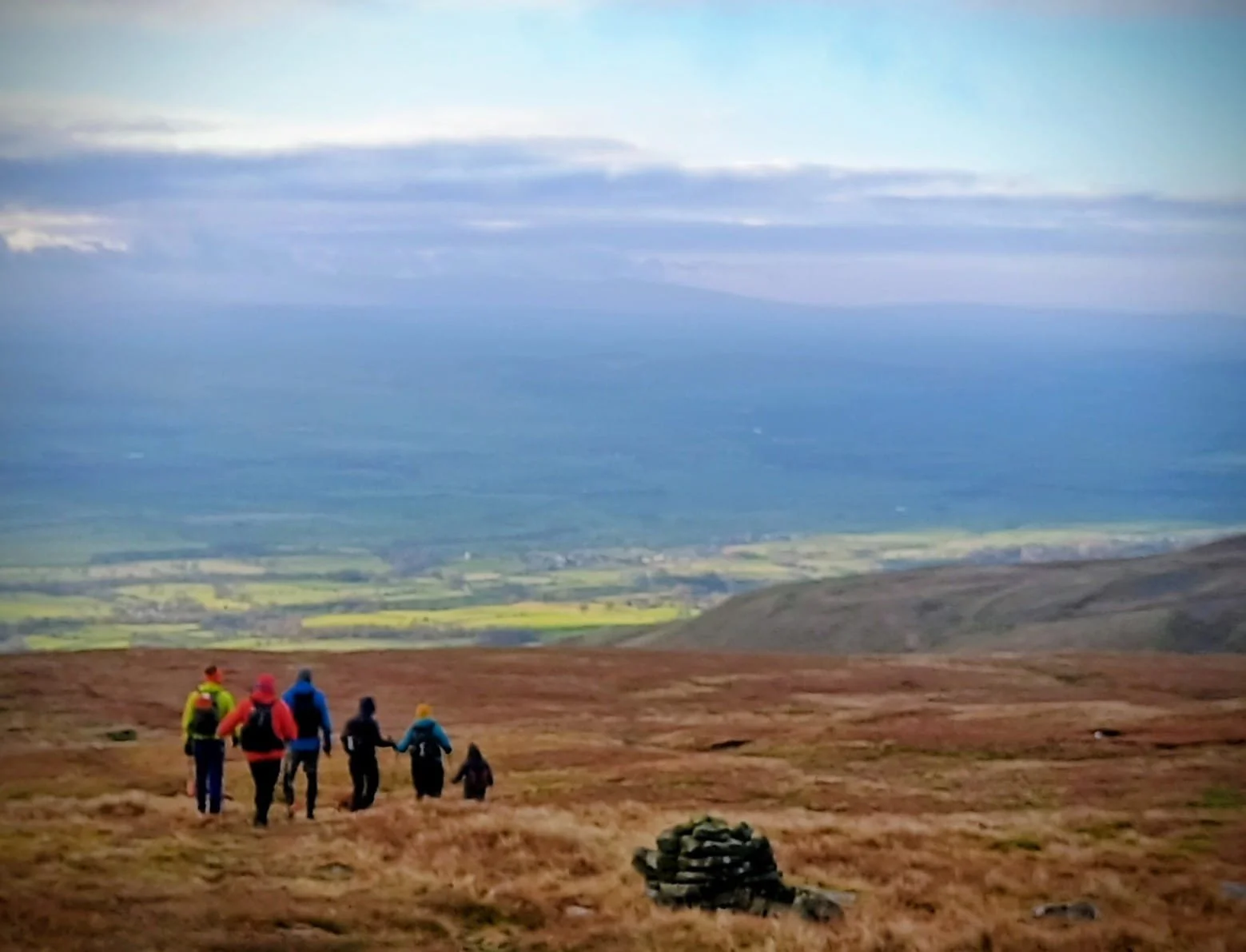 A group of hikers walking on a grassy hillside with a distant valley and mountains in the background.