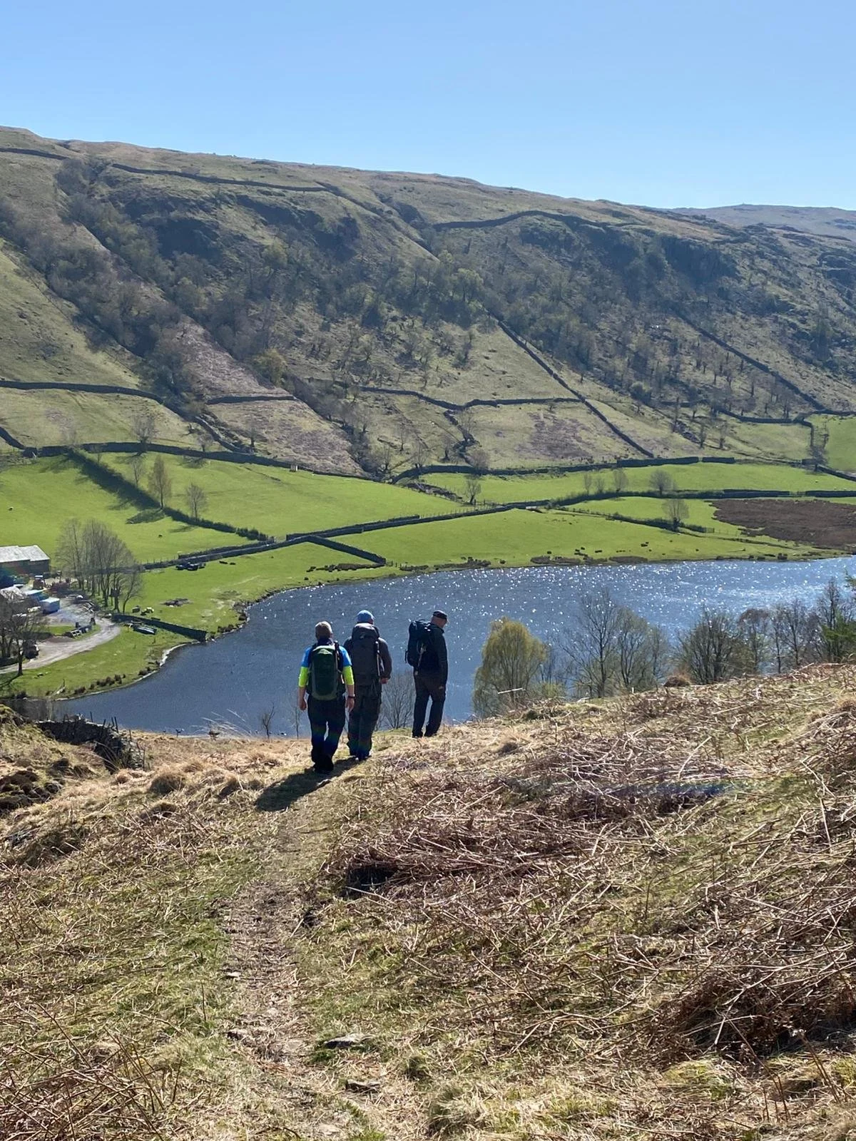 Three hikers with backpacks walking along a dirt trail overlooking a lake and green fields with a mountain in the background.