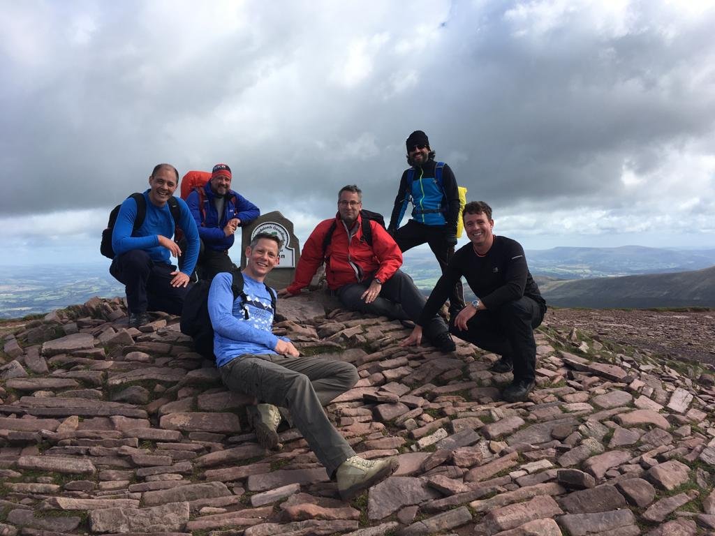 A group of six hikers on a rocky mountain top with cloudy skies and a panoramic view in the background.