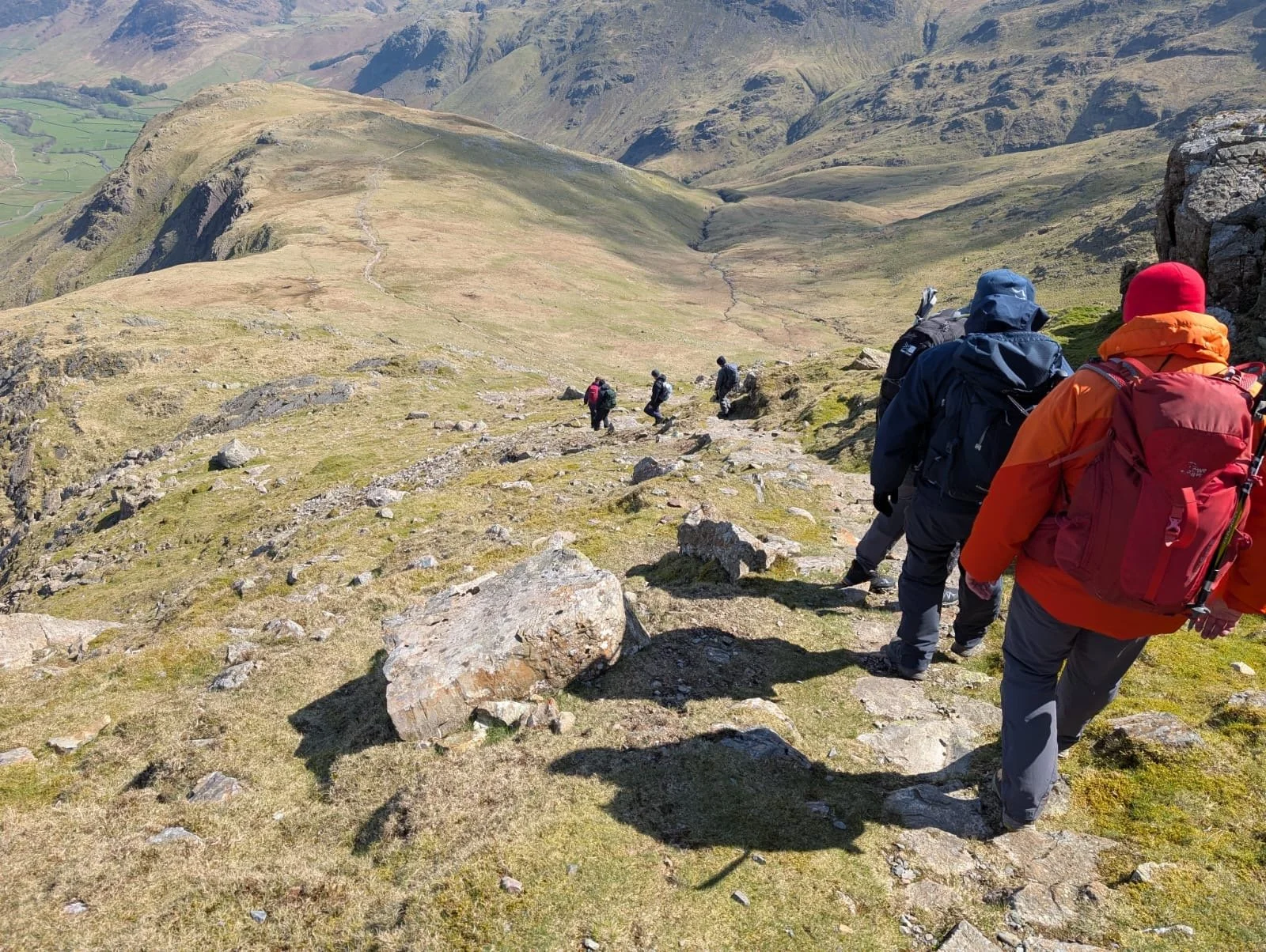 A group of hikers walking along a mountain trail in a scenic, hilly landscape with grassy patches and rocky terrain.