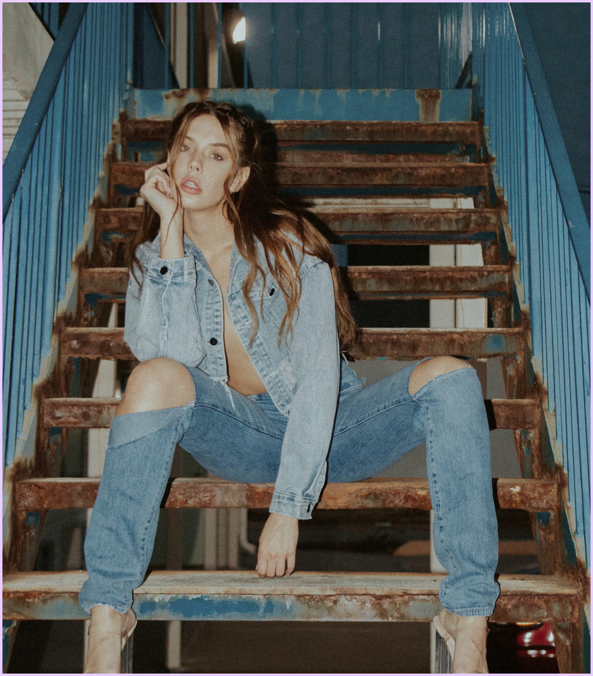 A young woman with long brown hair, wearing a light blue denim jacket and ripped jeans, poses sitting on old, rusty wooden stairs with blue railings, in an industrial or urban setting.