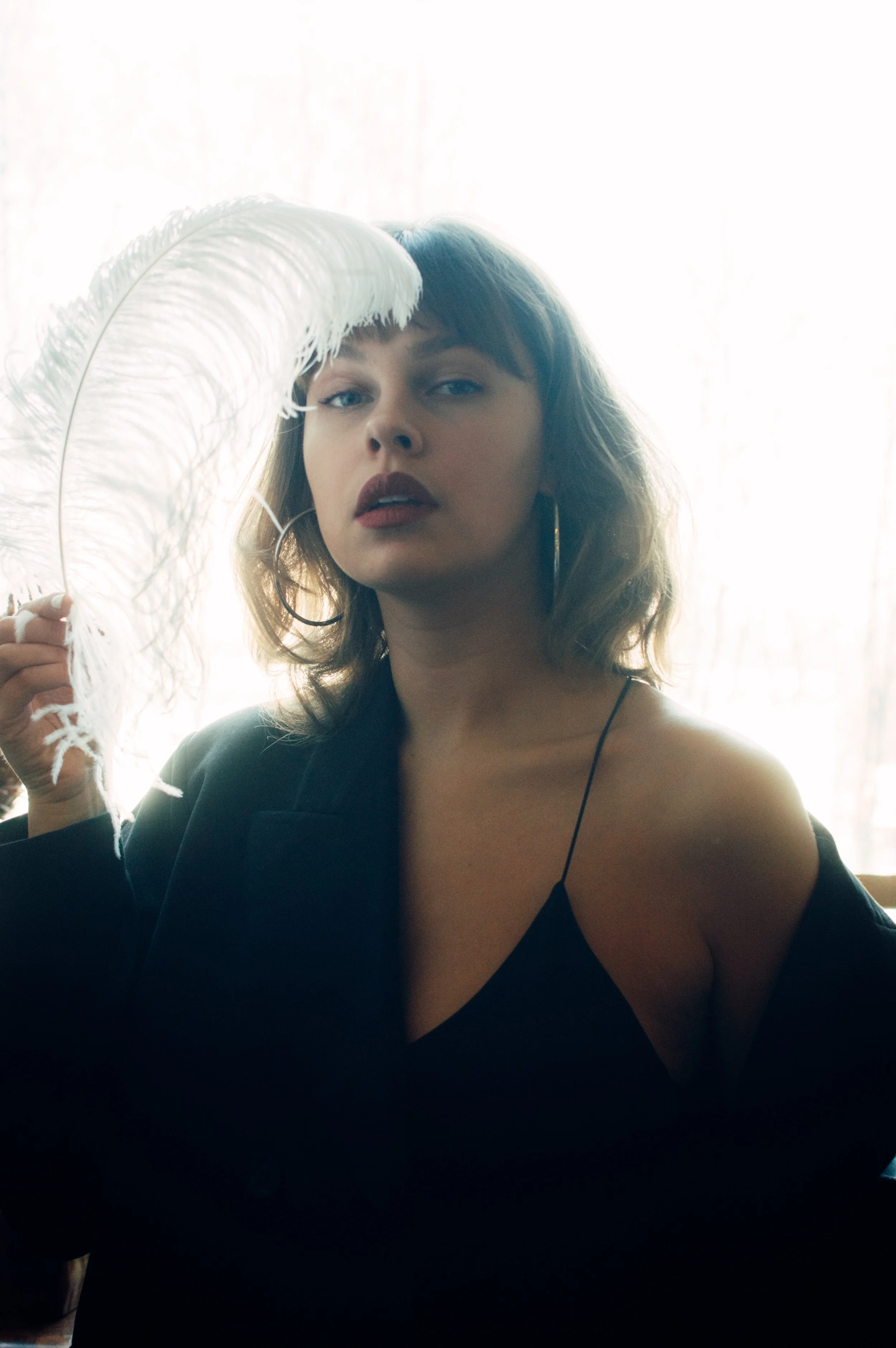 A woman with shoulder-length hair holding a large white feather quill in front of a bright window. She is wearing a black top with thin straps and large hoop earrings, looking at the camera with a neutral expression.