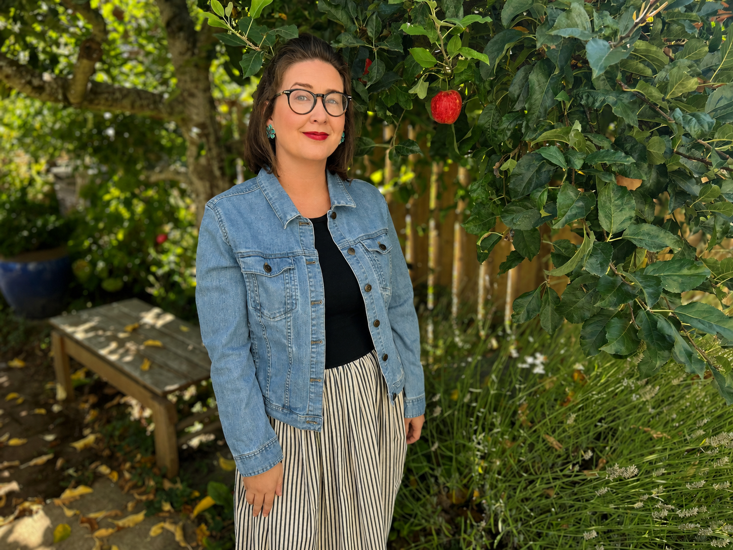 Emily Grosvenor with glasses and red lipstick standing outdoors next to a leafy tree with a red apple hanging from one of its branches.