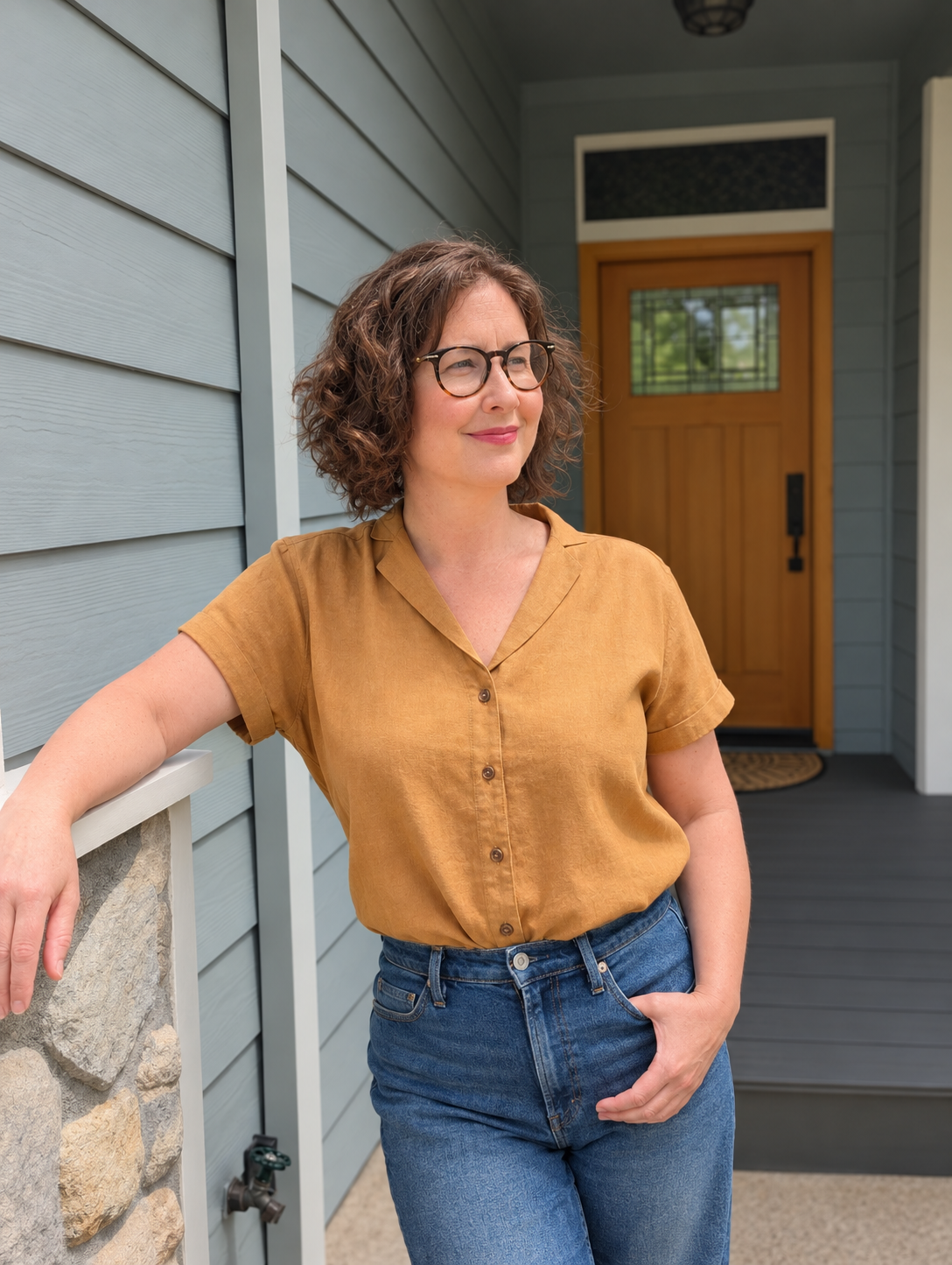 Emily Grosvenor with glasses and curly hair wearing a mustard yellow shirt and blue jeans, leaning against a stone and siding house exterior.