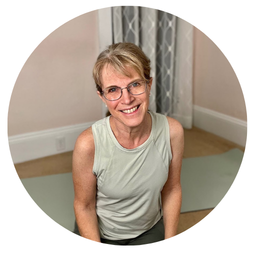 Pilates instructor Angie Floratos smiling woman with glasses and short blonde hair, wearing a sleeveless light green shirt, sitting in a room with beige walls and a window with curtains.