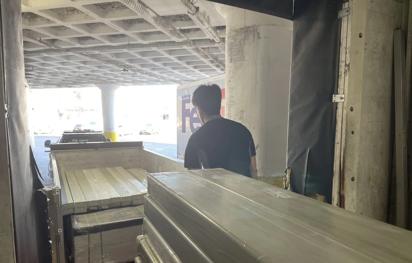 Person standing in a loading dock area, facing outside where cars are parked, with boxes and pallets in the foreground.