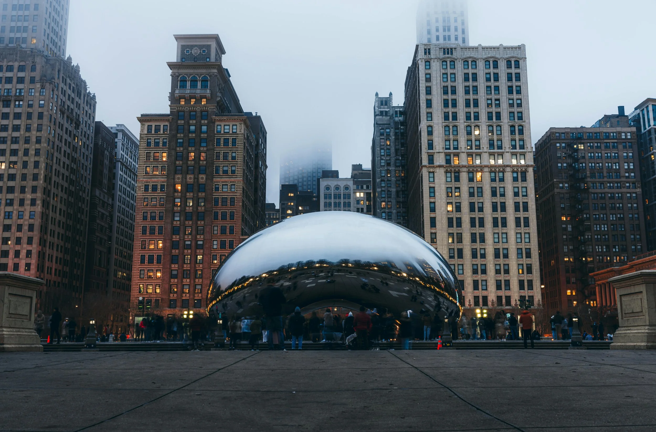 Nighttime view of a city square with tall buildings and the Cloud Gate sculpture (The Bean) reflecting the skyline and a crowd of people nearby.