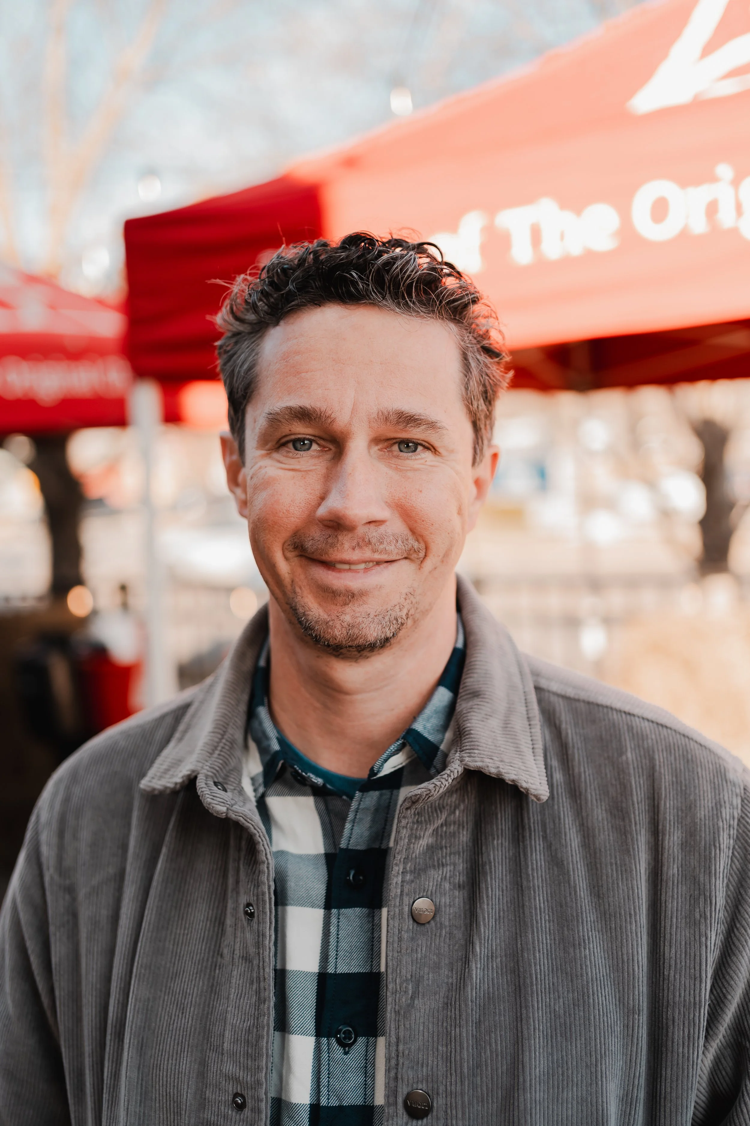 A man with short curly dark hair, blue eyes, and a light beard smiling outdoors with red awnings in the background.