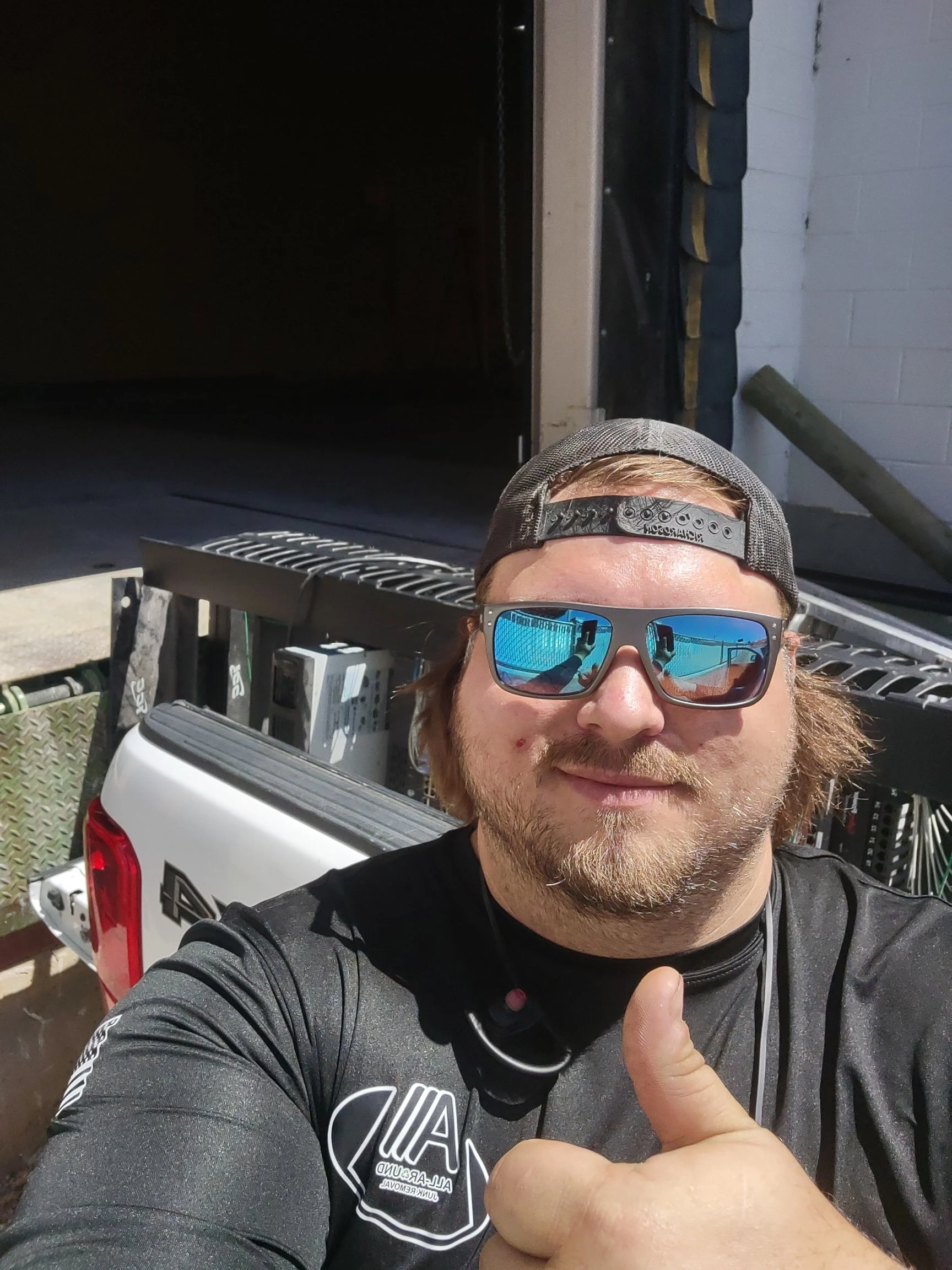 A man wearing sunglasses and a black long sleeve shirt with a logo on it is giving a thumbs up. He is also wearing a cap backward. He is taking a selfie with a vehicle and some tools and equipment in the background.