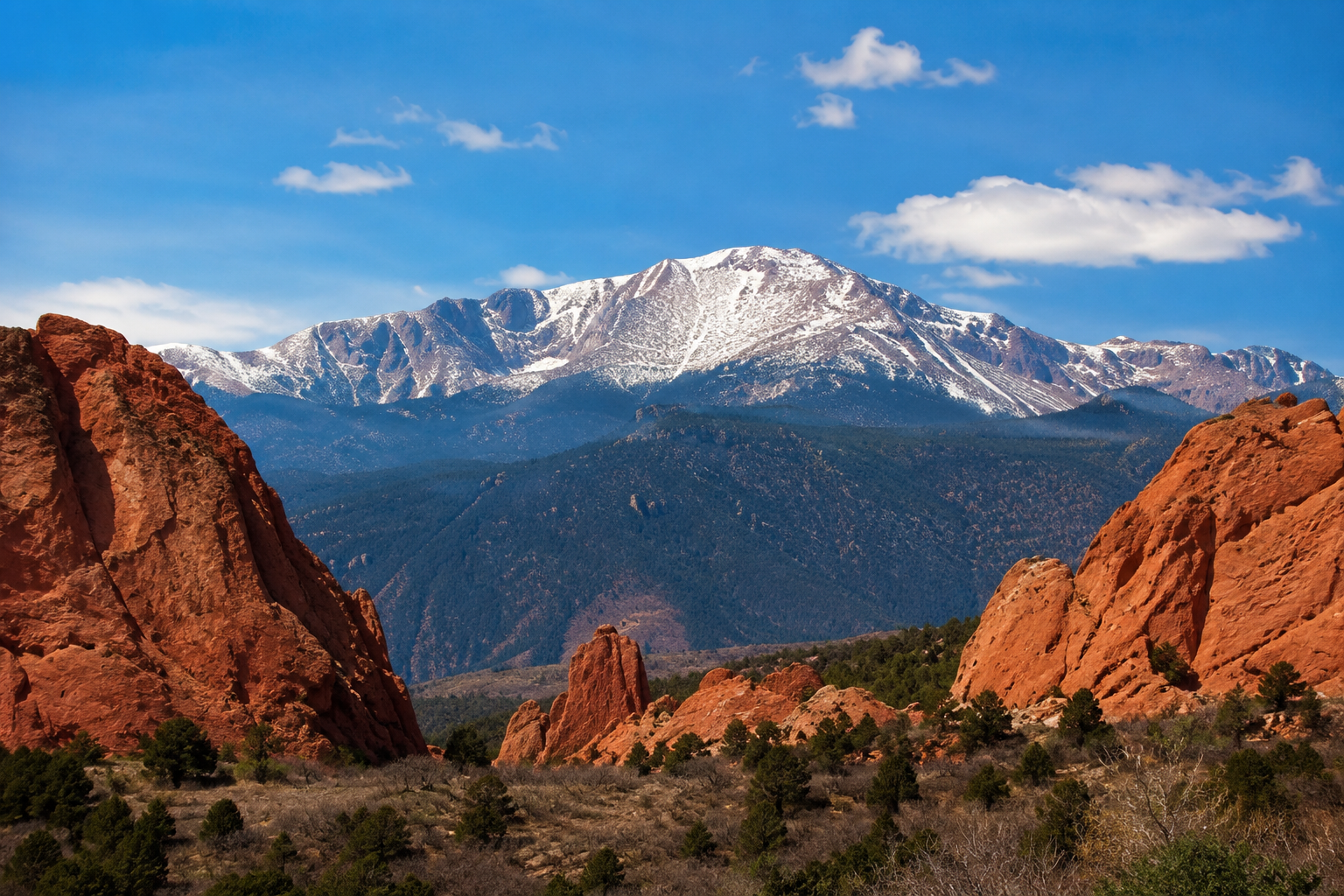 Snow-capped mountain in the background with red rock formations and green trees in the foreground under a blue sky with scattered clouds.