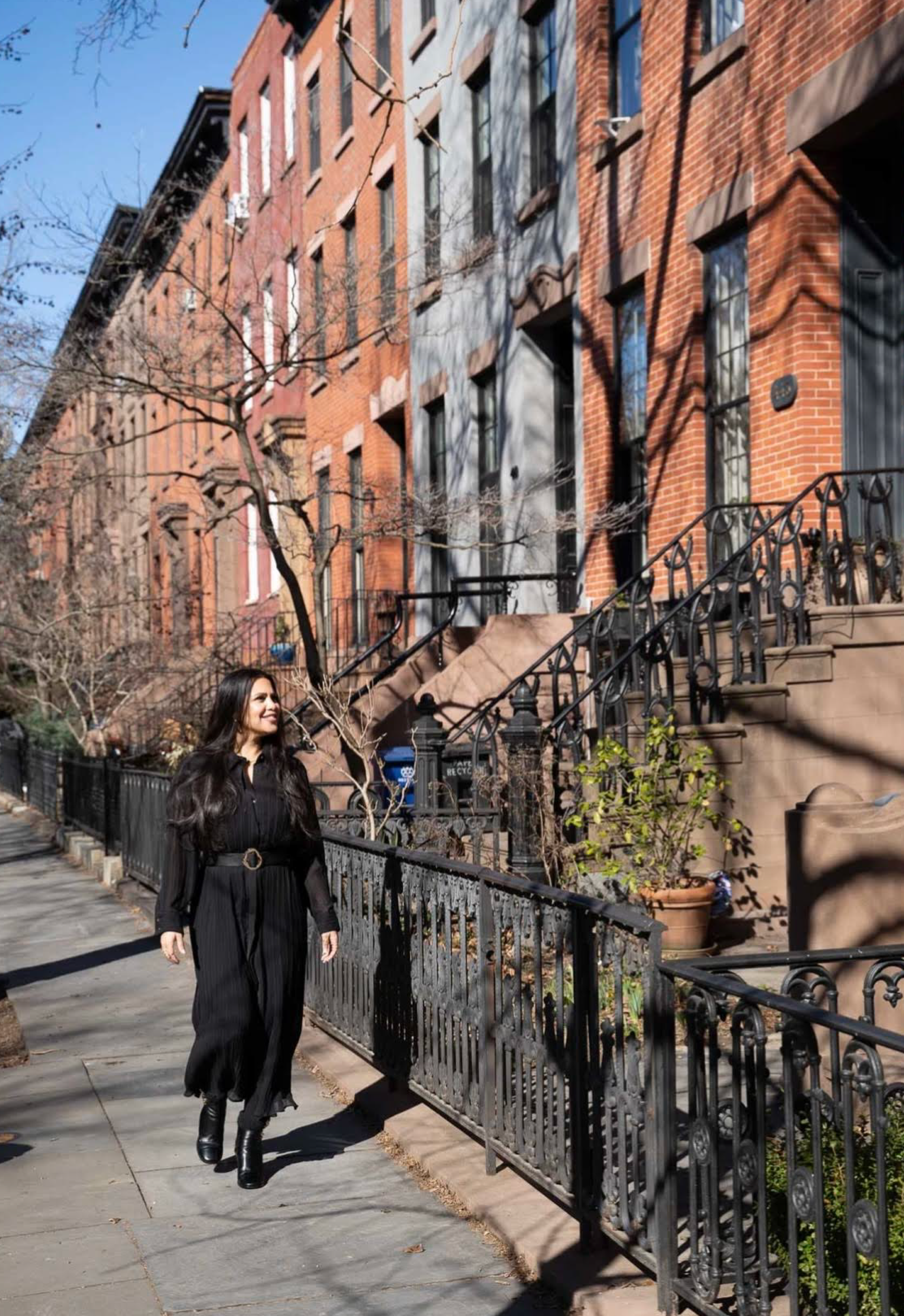 A woman with long dark hair wearing a black jumpsuit and black boots is walking on a city sidewalk. There are brownstone buildings with steps and iron railings signifying a neighborhood in New York City. Bare trees suggest it is winter.
