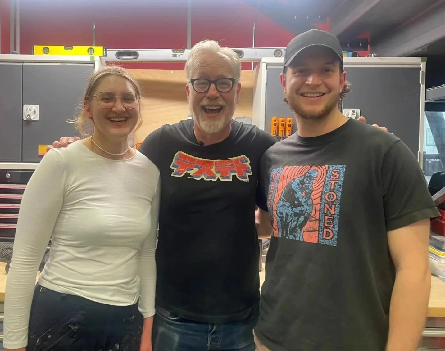 Three people smiling and posing together in a workshop or tool room. Two men and one woman, all casually dressed.