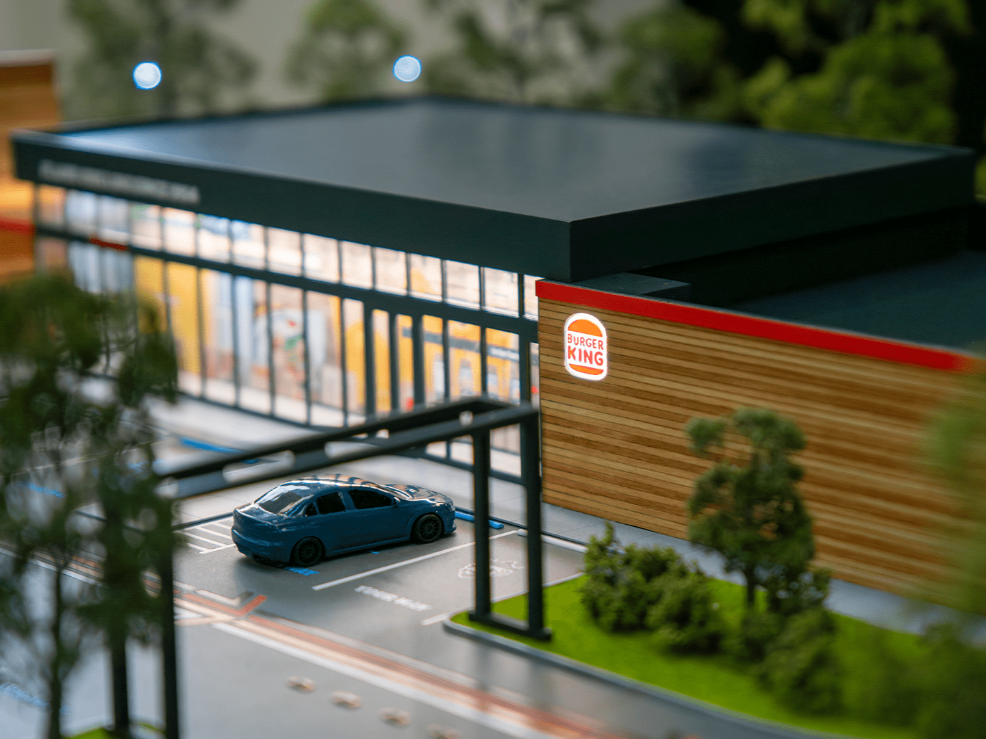 Miniature model of a Burger King restaurant with a parking lot, a blue car, trees, and surrounding greenery.