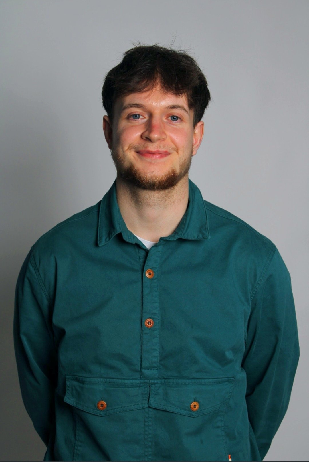 A young man with brown hair, blue eyes, and a light beard wearing a teal button-up shirt, standing against a plain gray background.