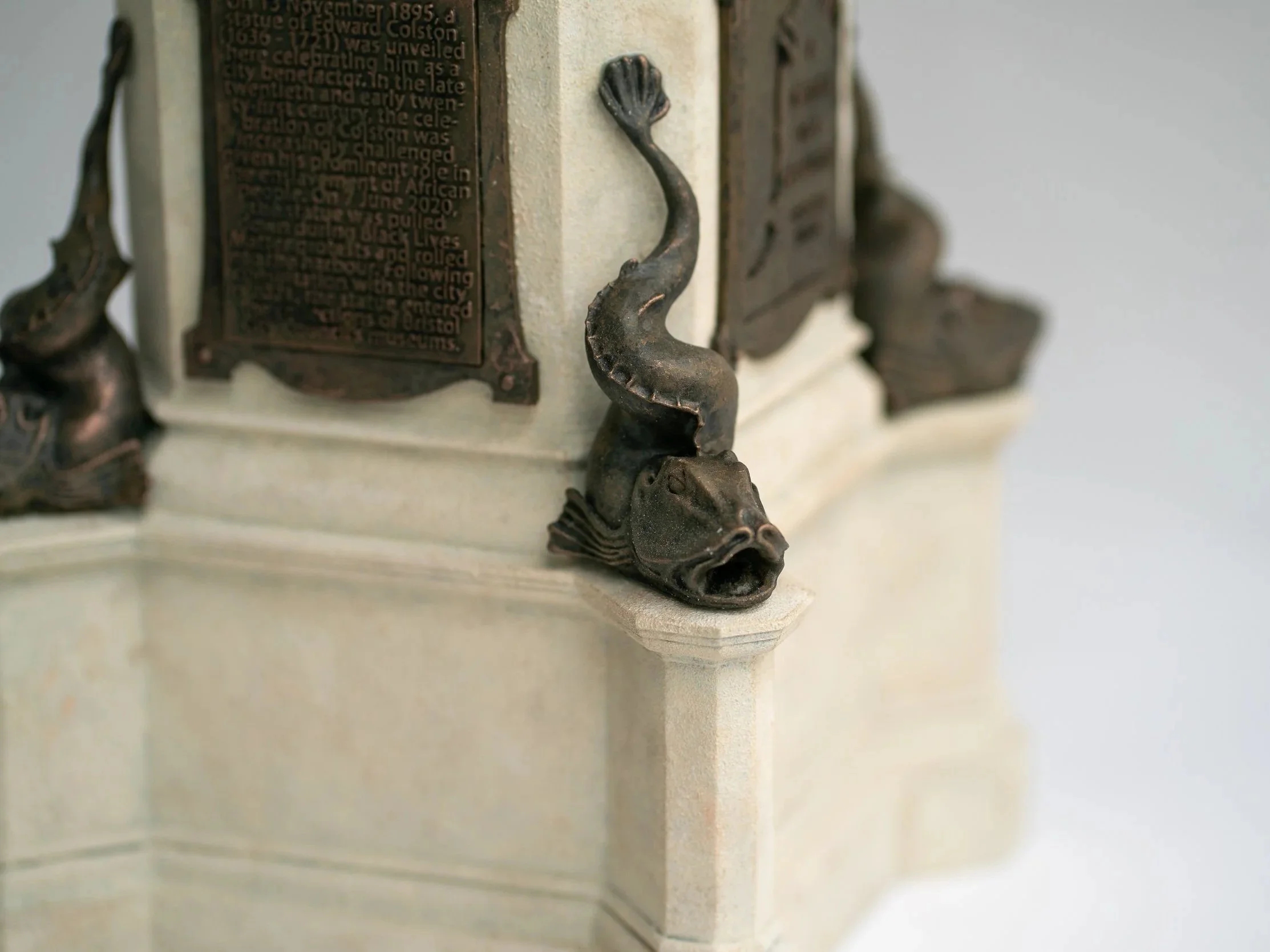 Close-up of a scale model of Bristol statue with a metal decorative element shaped like a fish attached to sand stone plinth.