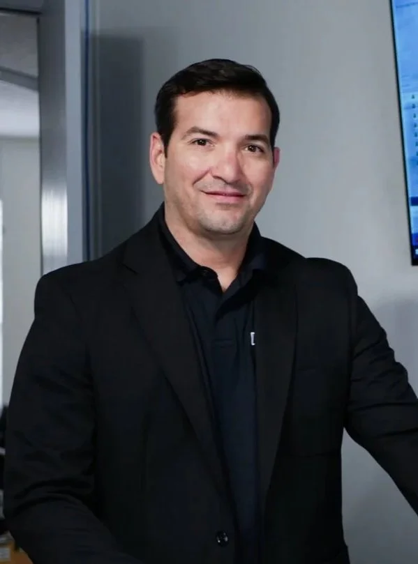 A man with dark hair in a black suit and collared shirt standing indoors near a monitor.
