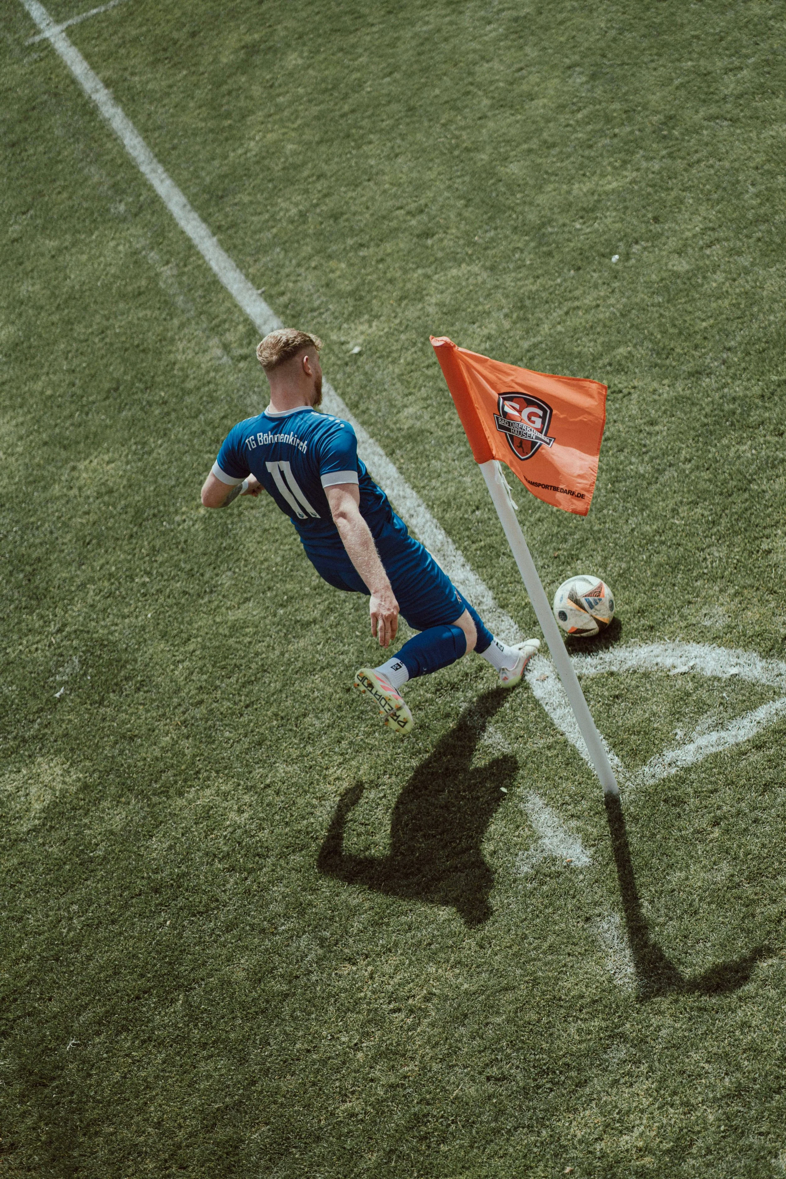 A white man in a blue uniform is shown on a green soccer pitch getting ready to kick a ball from the corner. There is a flag pole in the corner with an orange flag.