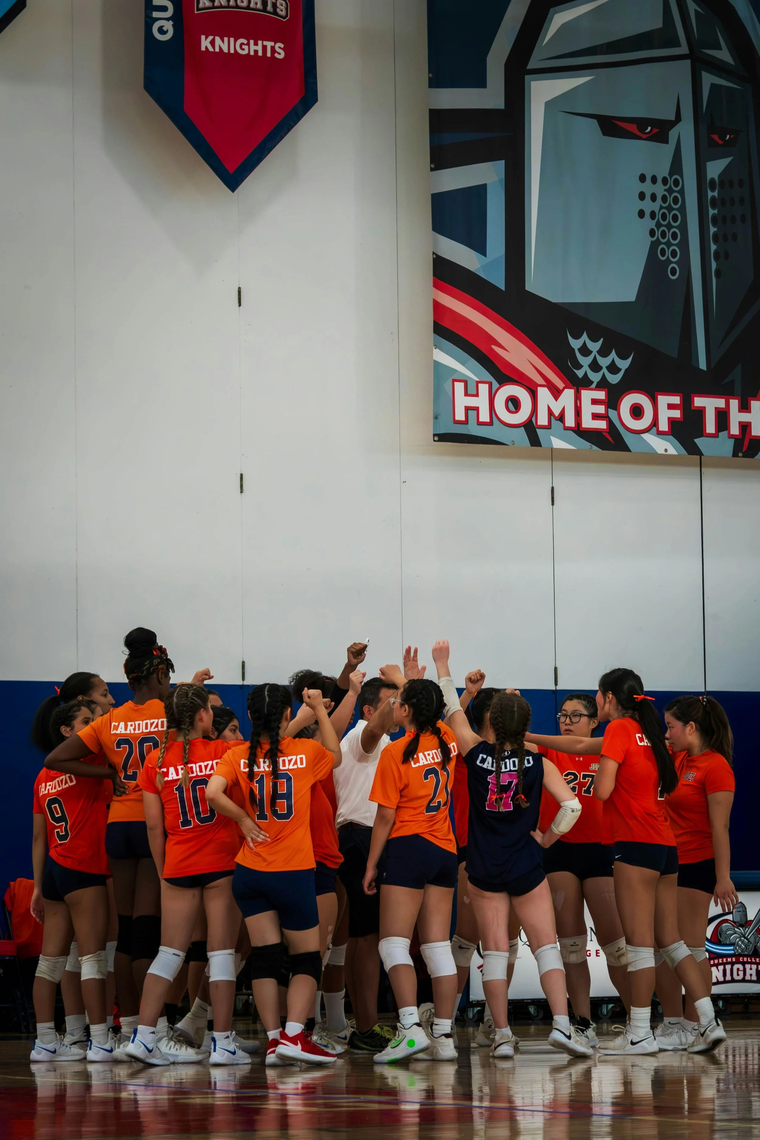 A girl's volleyball team wearing most orange jerseys is shown doing a team huddle in a gymnasium..