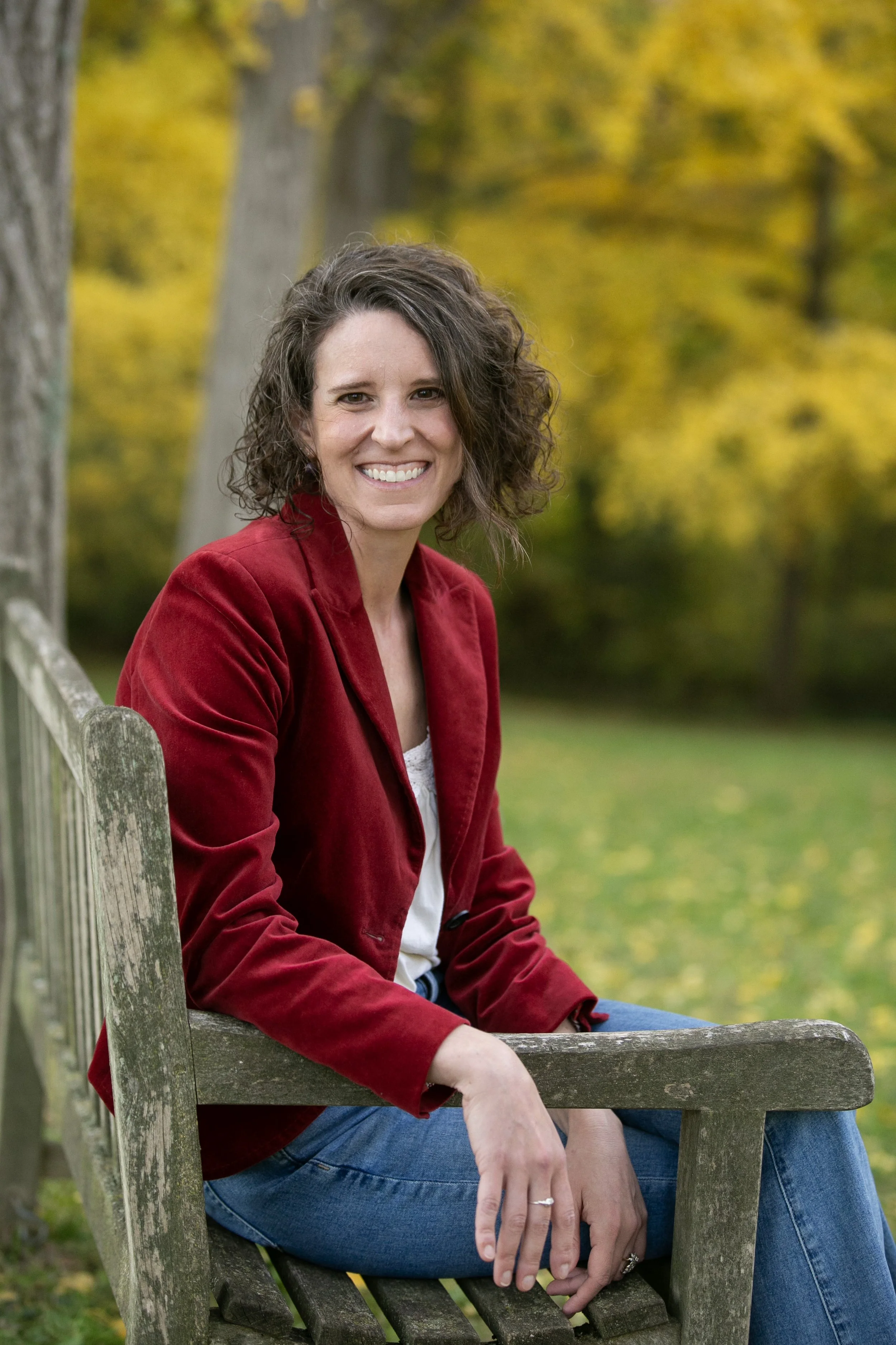 Jessica Peacock is shown sitting on a bench with yellow and green gingko trees in the background. She is a white woman with short curly brown hair and is wearing a red blazer and cream colored shirt.