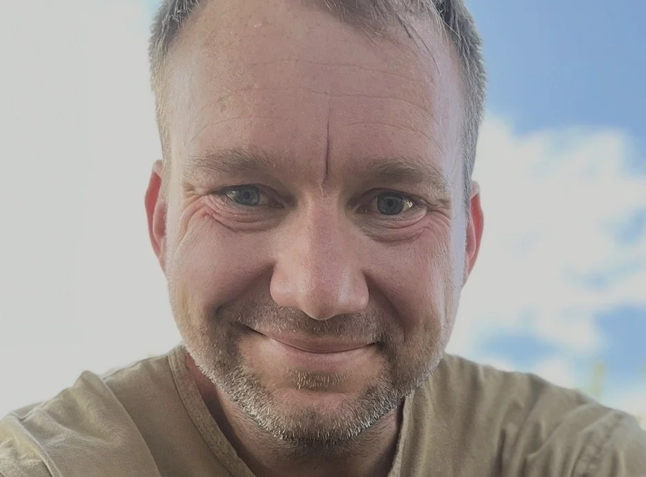 Close-up photo of a smiling man with short hair and a beard, outdoors with a blue sky and some clouds in the background.