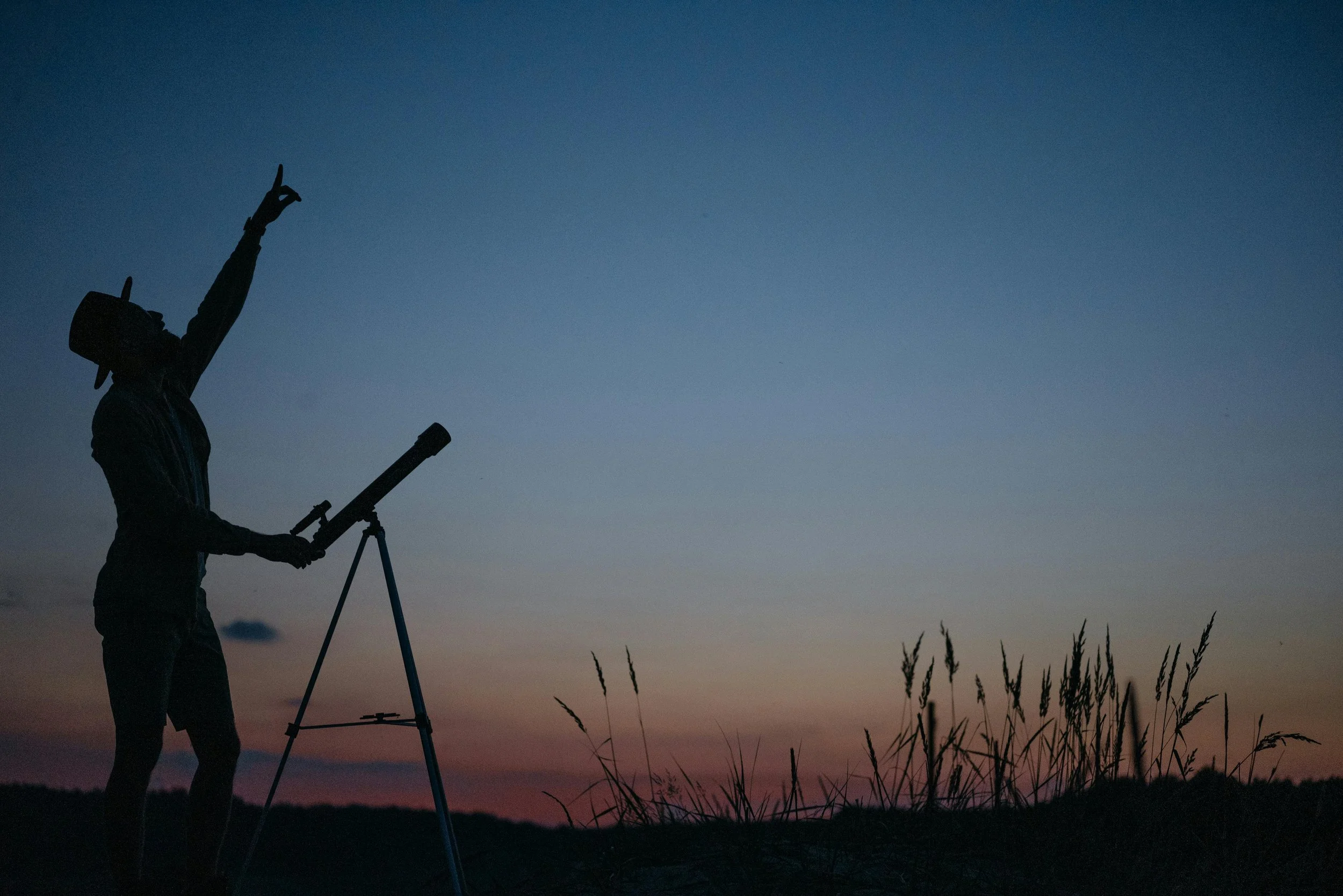 Person silhouetted against a twilight sky, using a telescope mounted on a tripod, with tall grass in the foreground.