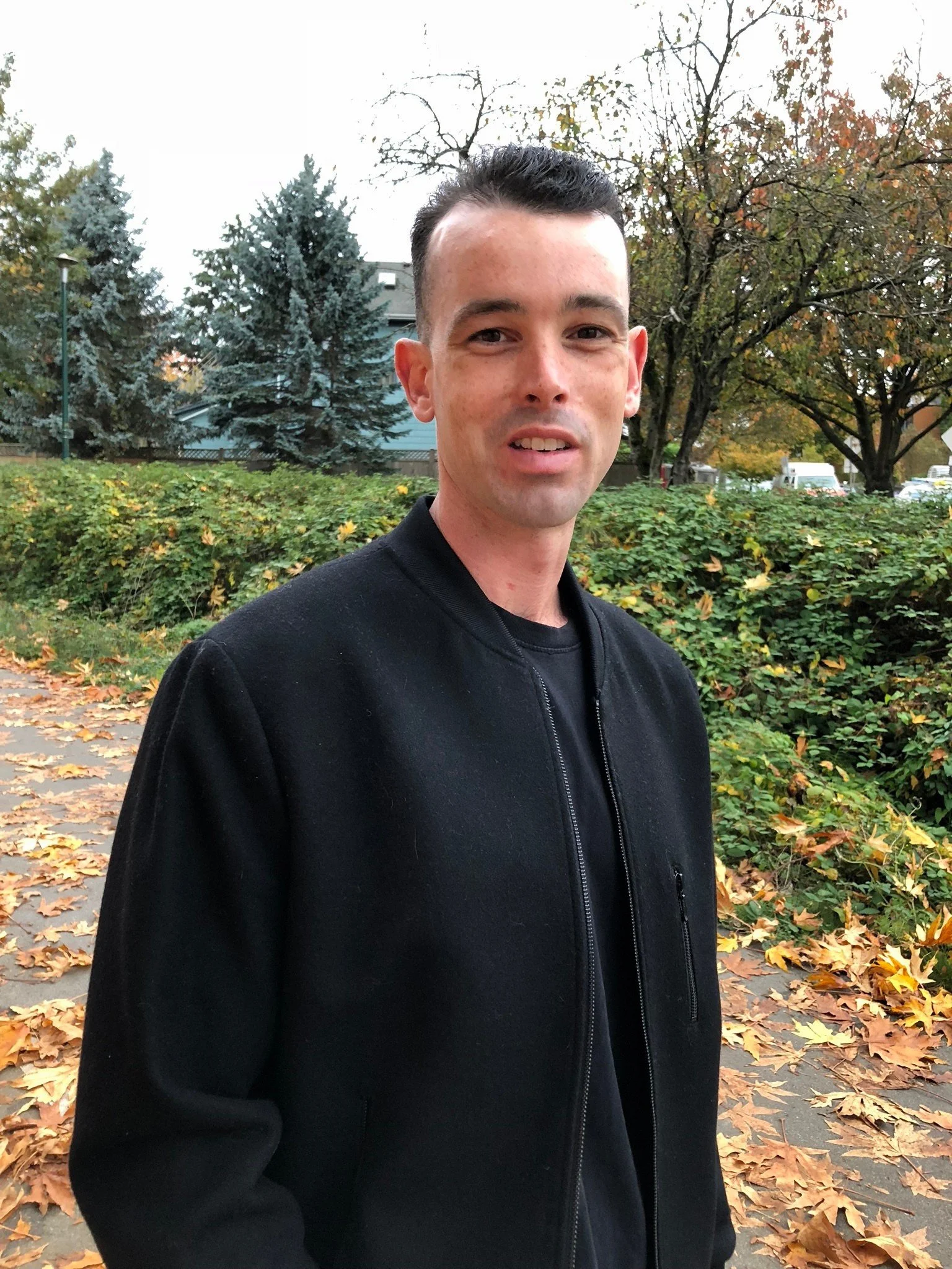A young man with short dark hair, wearing a black jacket, standing outdoors on a sidewalk covered in fallen autumn leaves, with trees and a house in the background.
