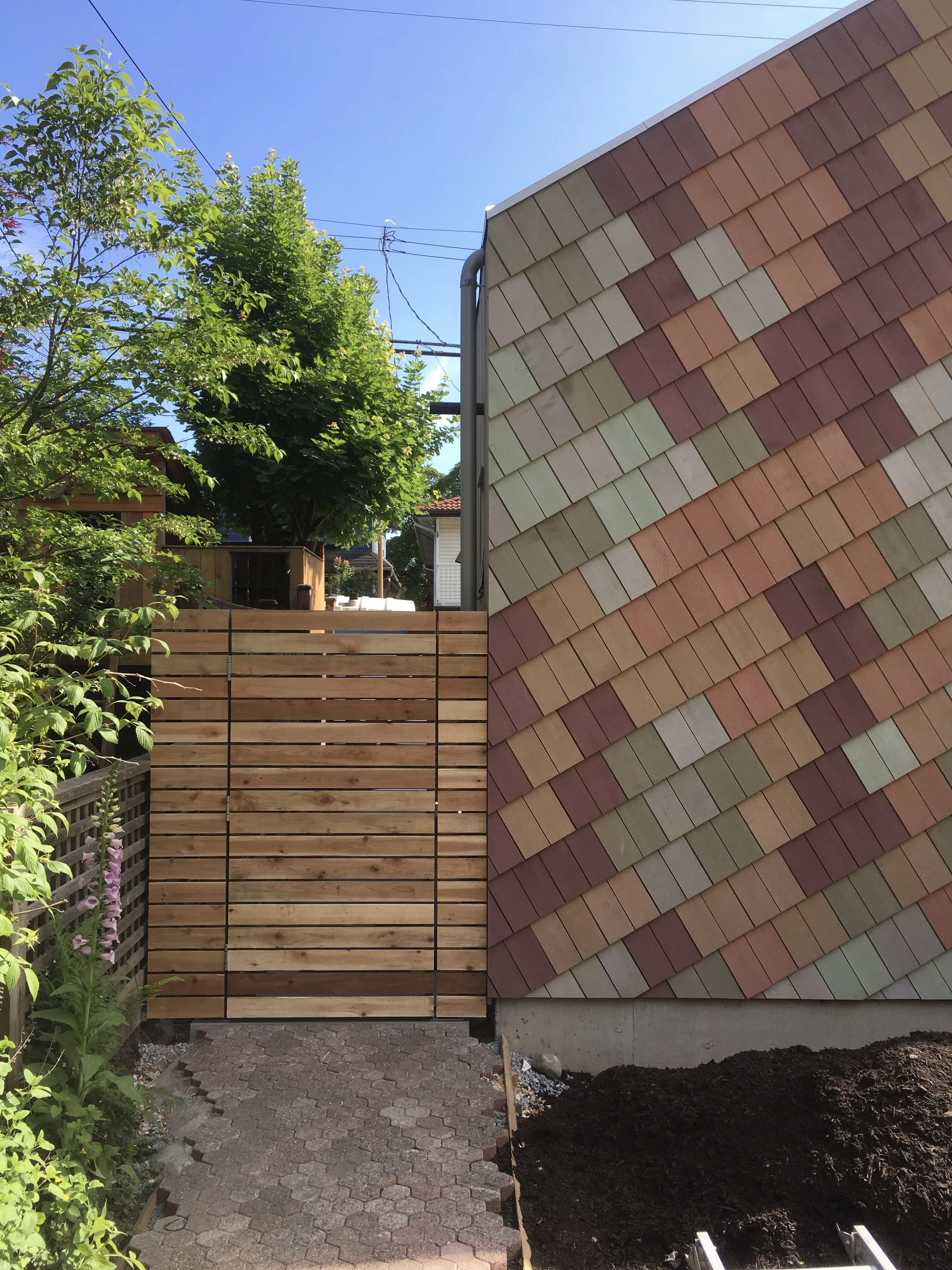 A backyard scene with a newly installed wooden fence and a decorative shingle wall. There are trees and plants along the sides, a brick pathway leading to the fence, and clear blue skies.