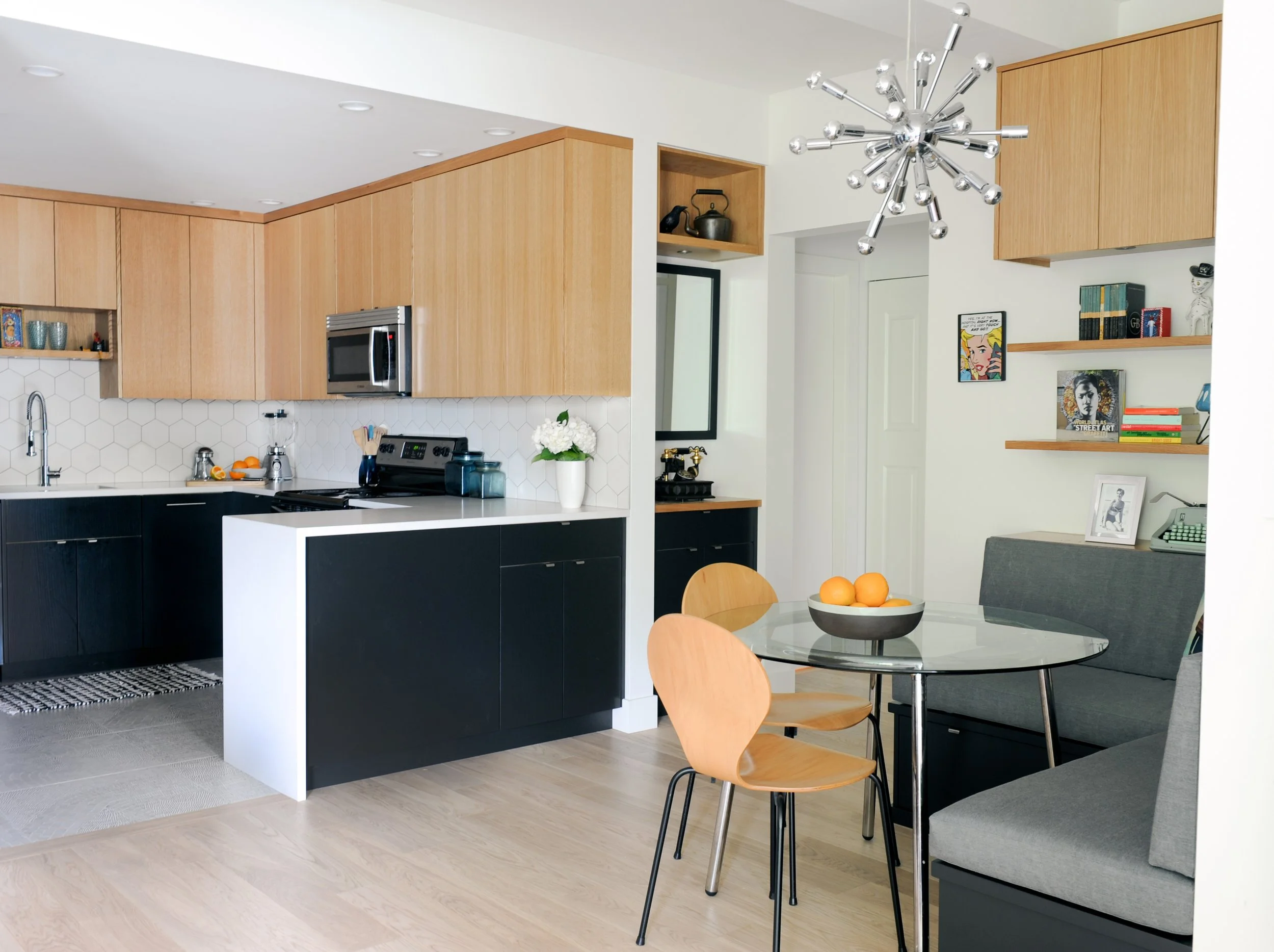 Modern open-concept kitchen and dining area with wooden cabinetry, black and white accents, a round glass table, wooden and upholstered chairs, decorative shelves with books and art, and a contemporary chandelier.