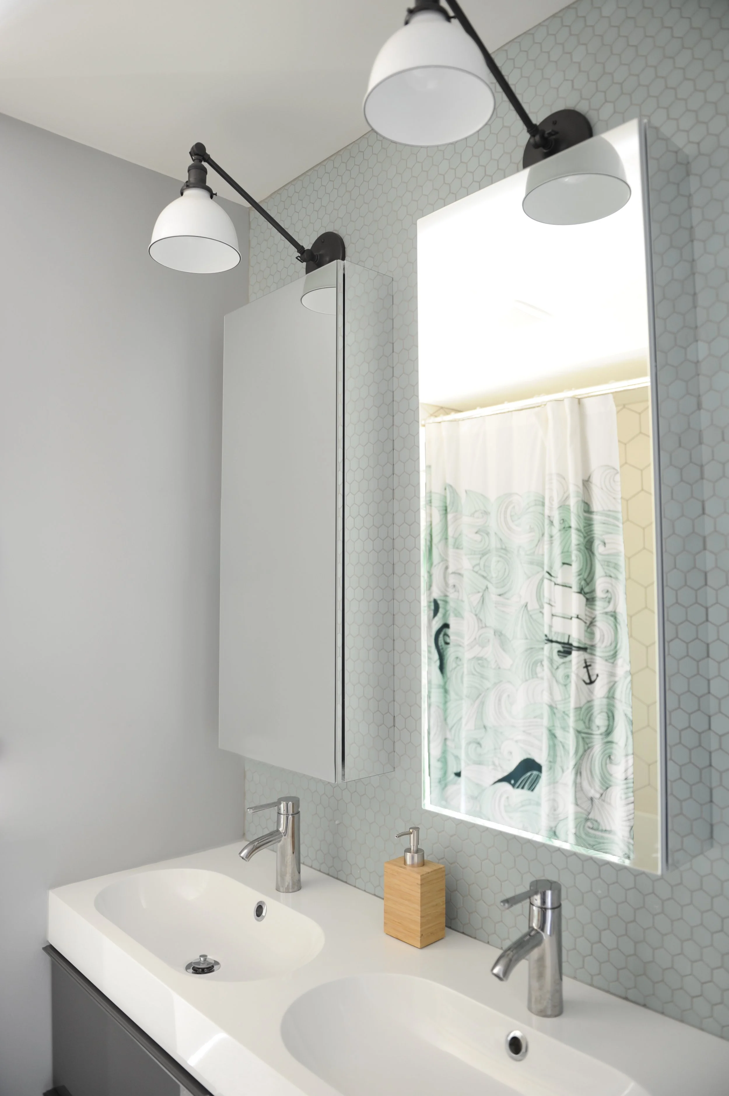 Bathroom vanity with double sinks, modern faucets, a wood tissue box, and a large mirror. The wall behind features light-colored hexagonal tiles, and a shower curtain with a wave pattern is visible in the mirror's reflection.